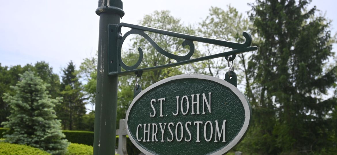 Green circular street sign reading 'St John Chrysostom' mounted on an ornate dark-green post with trees in the background.