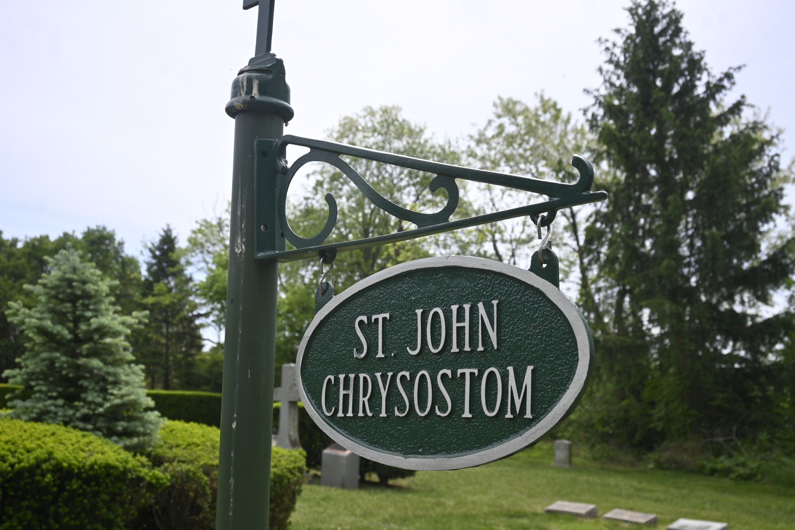 Green circular street sign reading 'St John Chrysostom' mounted on an ornate dark-green post with trees in the background.