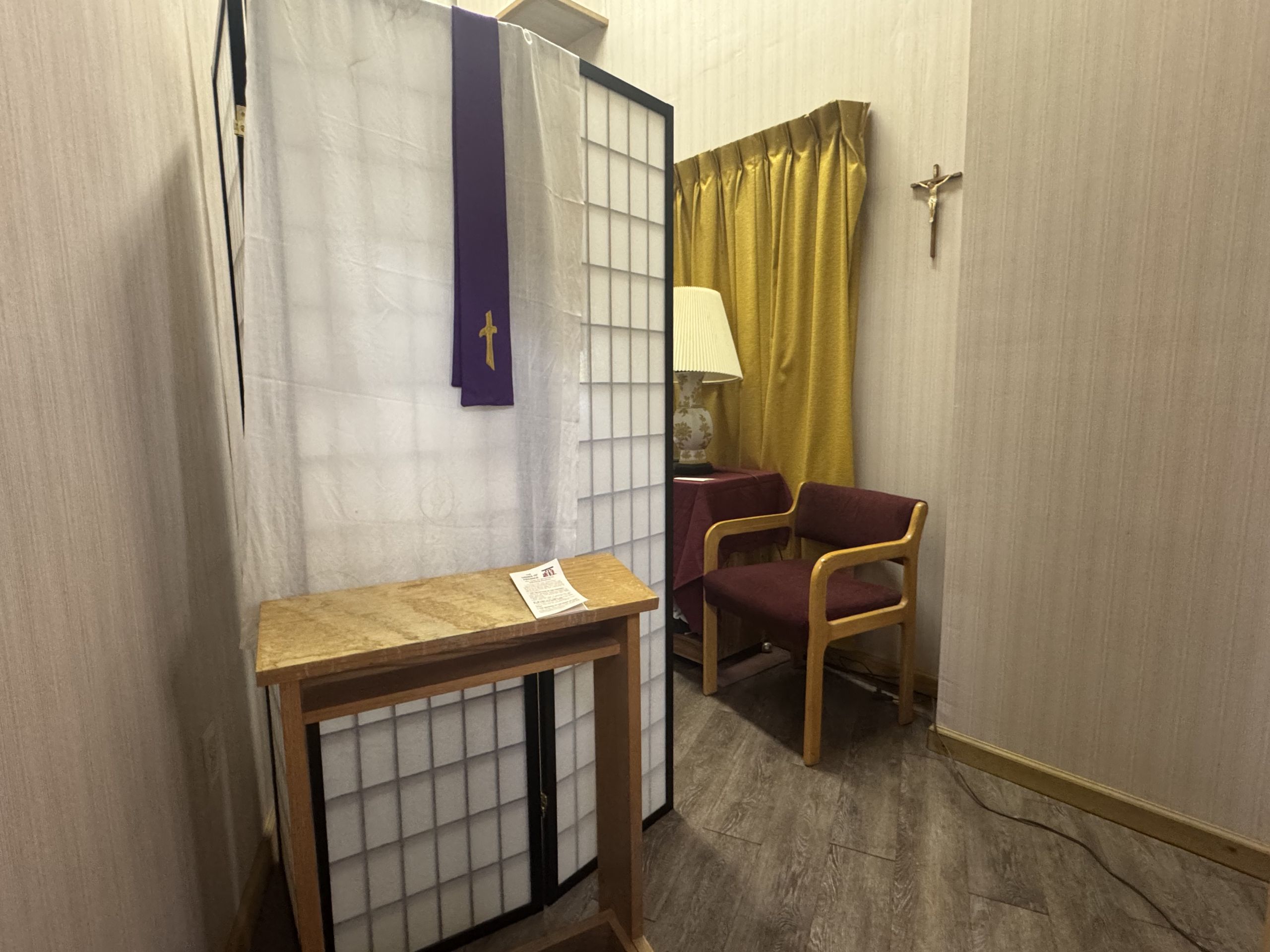 Interior of a small prayer or counseling room with a folding screen, a purple liturgical stole draped over it, and a wooden table with a flyer. A gold curtain and a couple of wooden chairs are in the background, and a crucifix hangs on the wall.