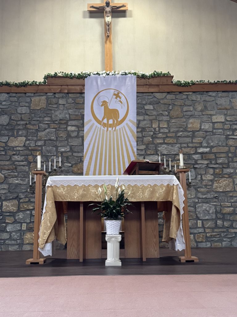 Church altar with a crucifix, floral garland, and a white banner showing a gold ram emblem and sunburst behind it.