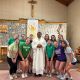 Twelve girls in colorful shirts pose with a smiling priest in white robes inside a church, standing in front of a stone altar with a crucifix.