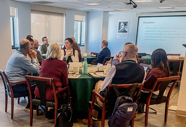 People seated around a conference table during a discussion, with a projection screen showing 'Discussion' in the room.