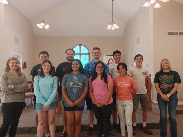 Group of teenagers posing for a photo in a church hall, smiling and standing on steps in front of a round window and pews.