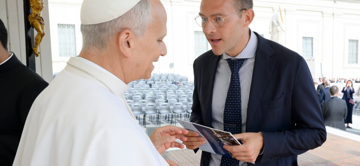Religious figure in white robes shakes hands with a man in a suit while sharing a pamphlet at an outdoor event with rows of chairs in the background.