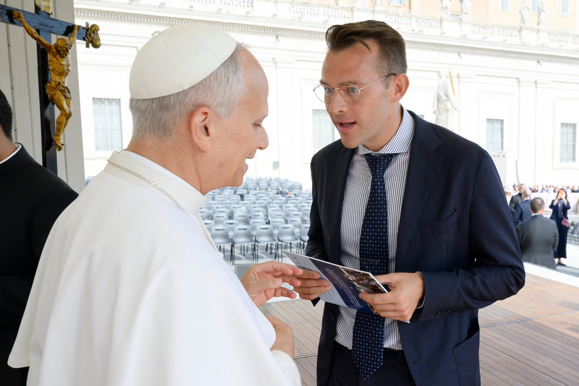 Religious figure in white robes shakes hands with a man in a suit while sharing a pamphlet at an outdoor event with rows of chairs in the background.