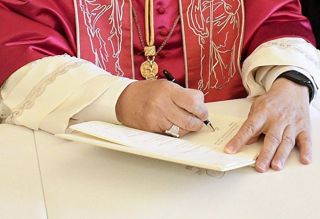 Clergy member in red liturgical vestments signs a document at a table, with a gold pendant visible around the neck.