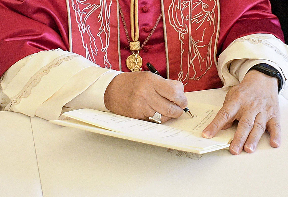 Clergy member in red liturgical vestments signs a document at a table, with a gold pendant visible around the neck.