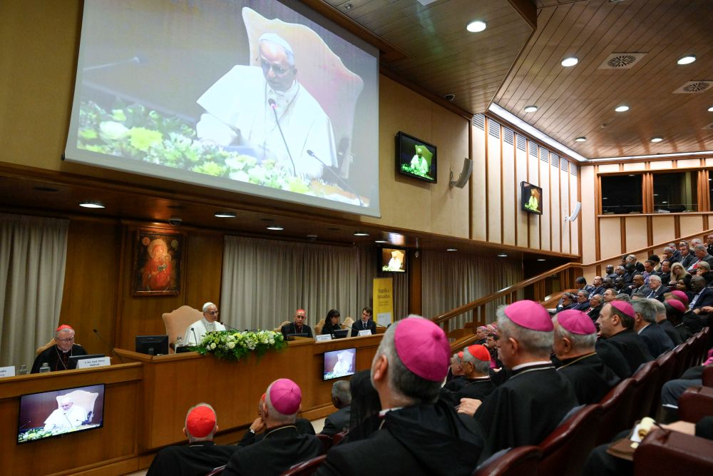 Religious leaders seated at a long conference table in a wood-paneled hall, with a large screen showing the speaker at the podium and floral arrangements in front.