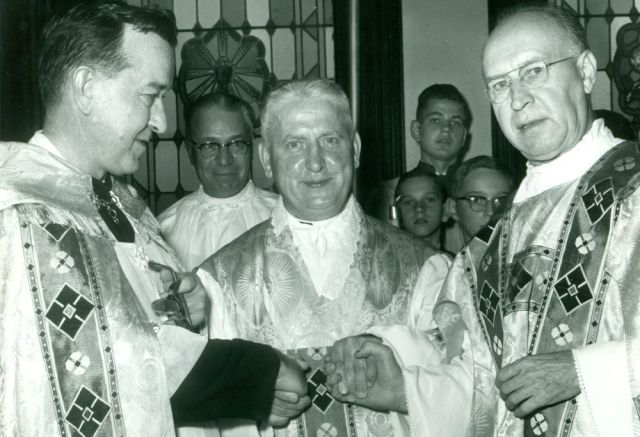 Three clergy in ornate vestments shake hands at a church ceremony, with a choir visible in the background.