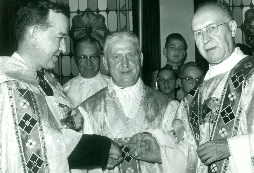 Three clergy in ornate vestments shake hands at a church ceremony, with a choir visible in the background.