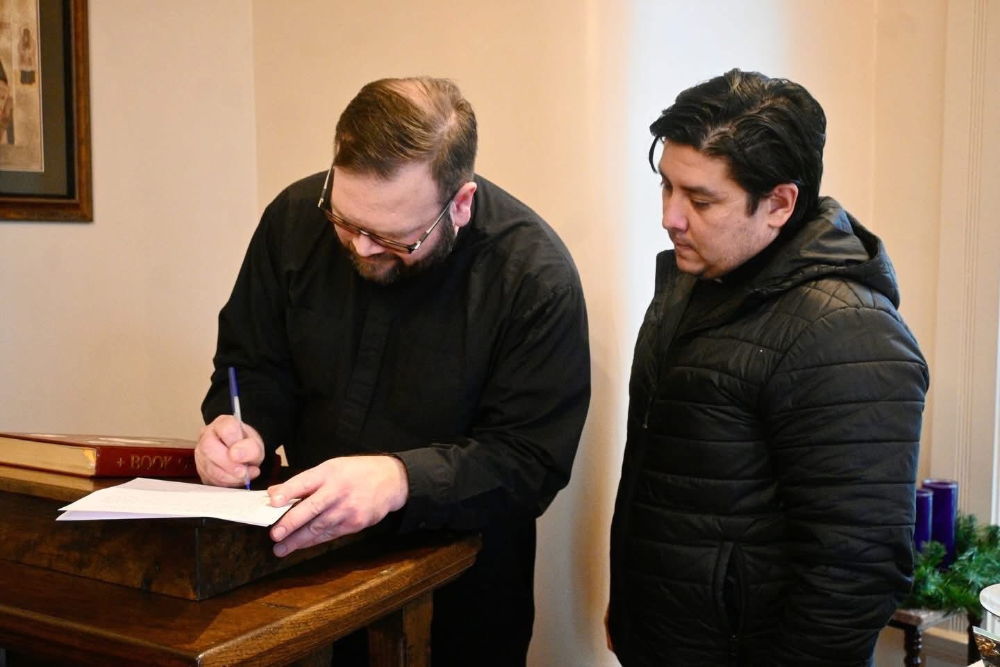 Man with glasses signs a document on a wooden lectern while another man watches.