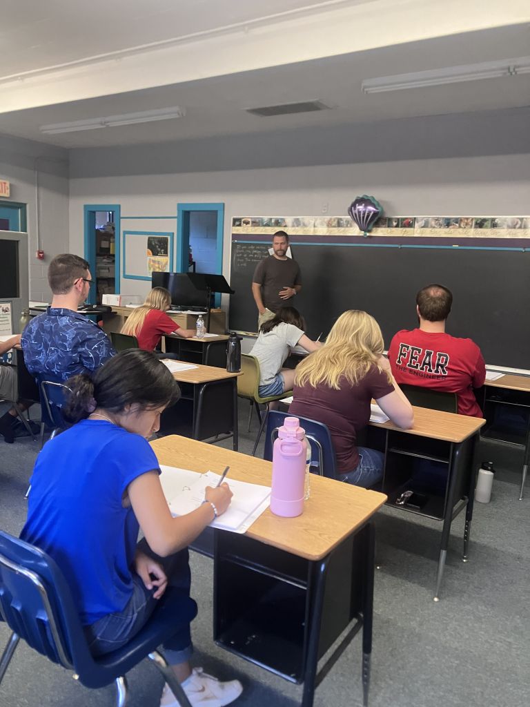 Classroom scene with students seated at desks, a teacher standing by the chalkboard at the front of the room.