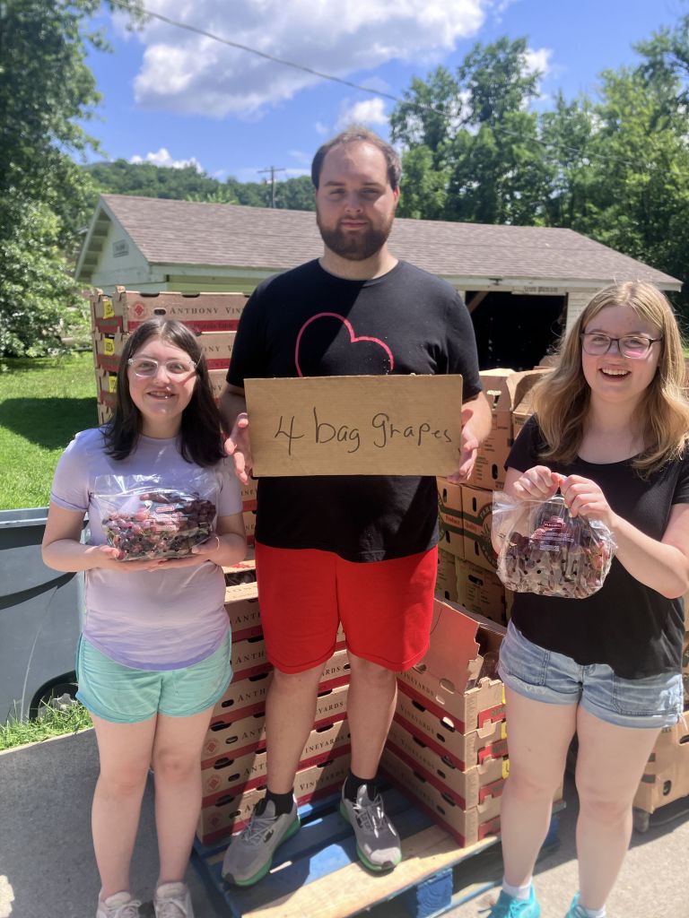Three smiling people standing outdoors with stacked grape boxes; the man in the center holds a cardboard sign reading '4 bag grapes'.