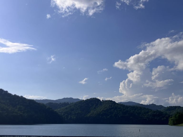 Calm lake with tree-covered hills in the distance under a blue sky with white clouds.