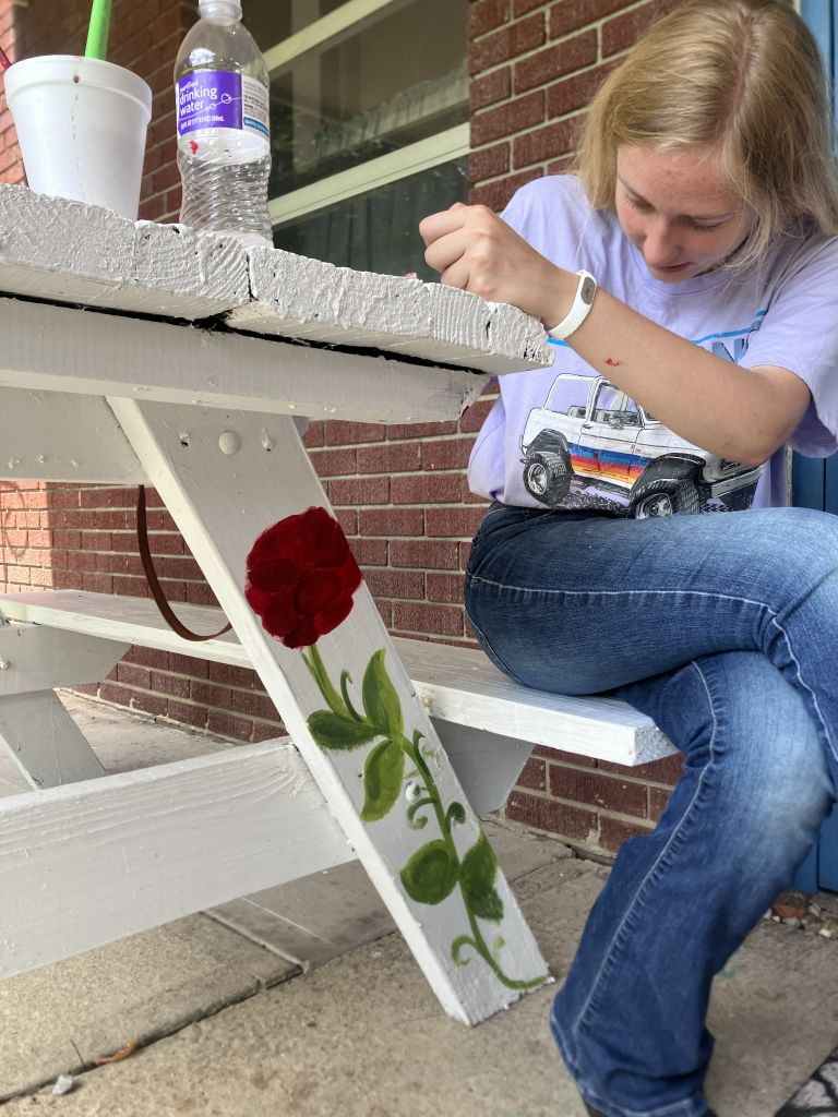 Girl painting a white wooden railing on a porch, with a bottle and cup nearby and a red brick wall in the background.