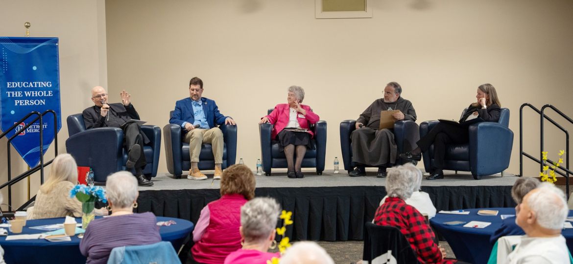 Five panelists seated in blue chairs on a stage, talking to an audience in a hall; banner on left reads 'Educating the Whole Person'