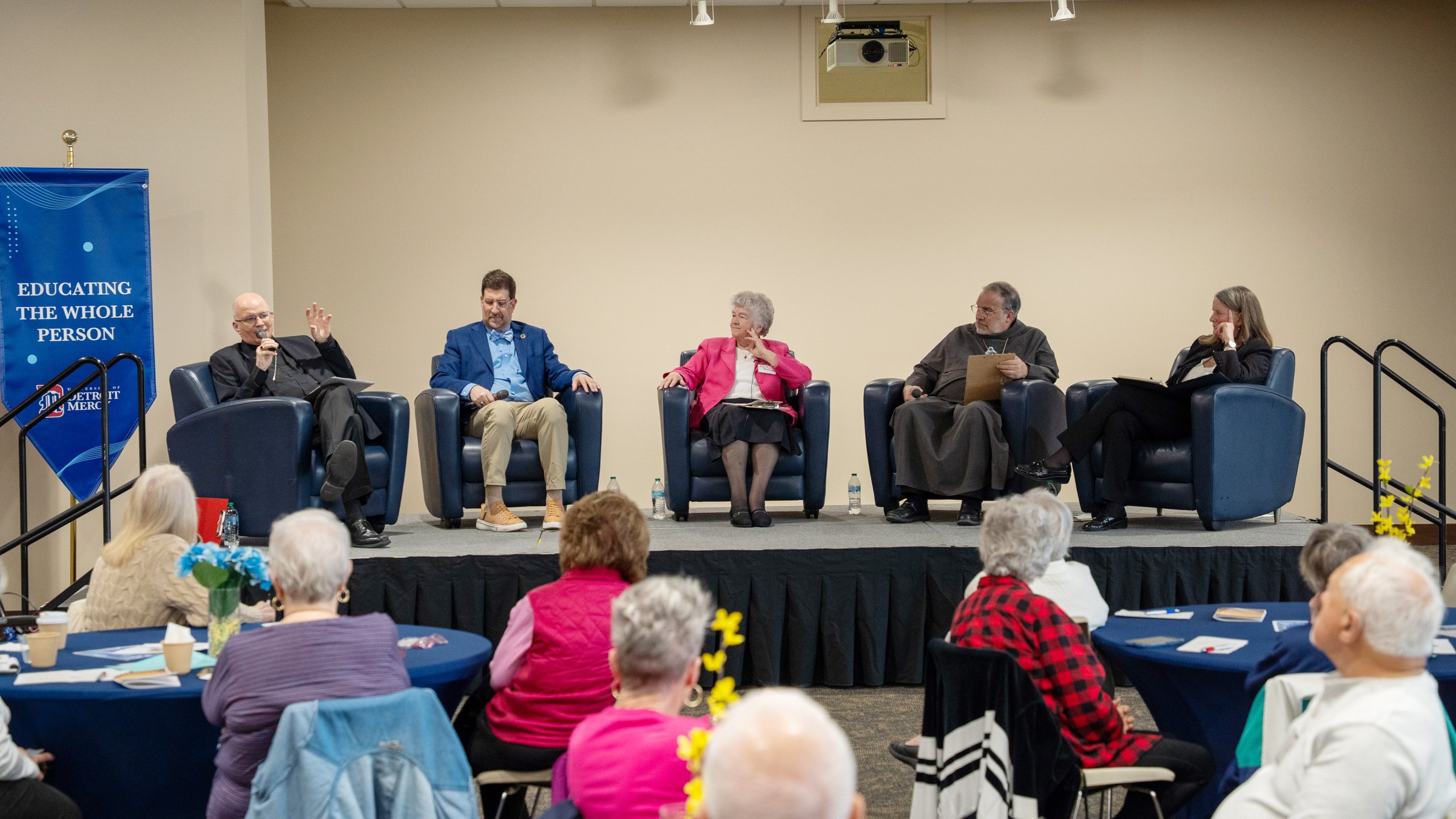 Five panelists seated in blue chairs on a stage, talking to an audience in a hall; banner on left reads 'Educating the Whole Person'