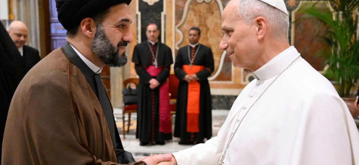 Pope Francis shakes hands with a Muslim cleric in brown robes and black turban in a ceremonial hall, with officials in the background.