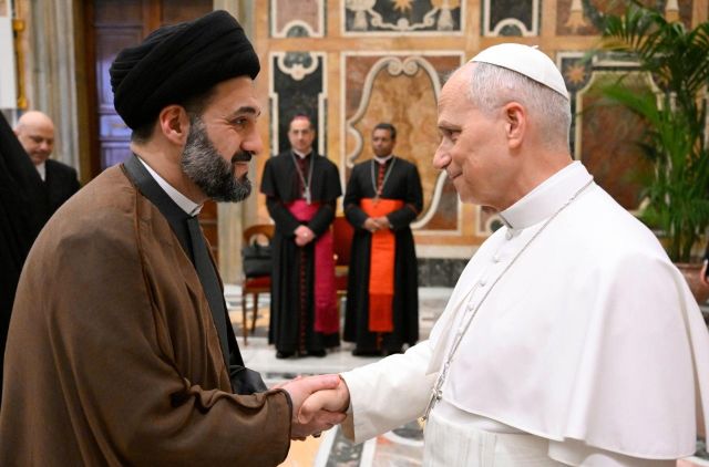 Pope Francis shakes hands with a Muslim cleric in brown robes and black turban in a ceremonial hall, with officials in the background.