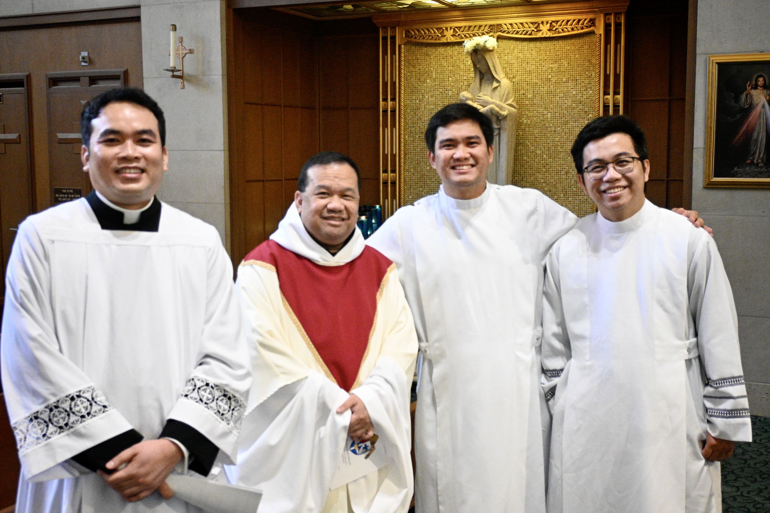 Four clergy members in white robes pose together inside a church, smiling for the camera.