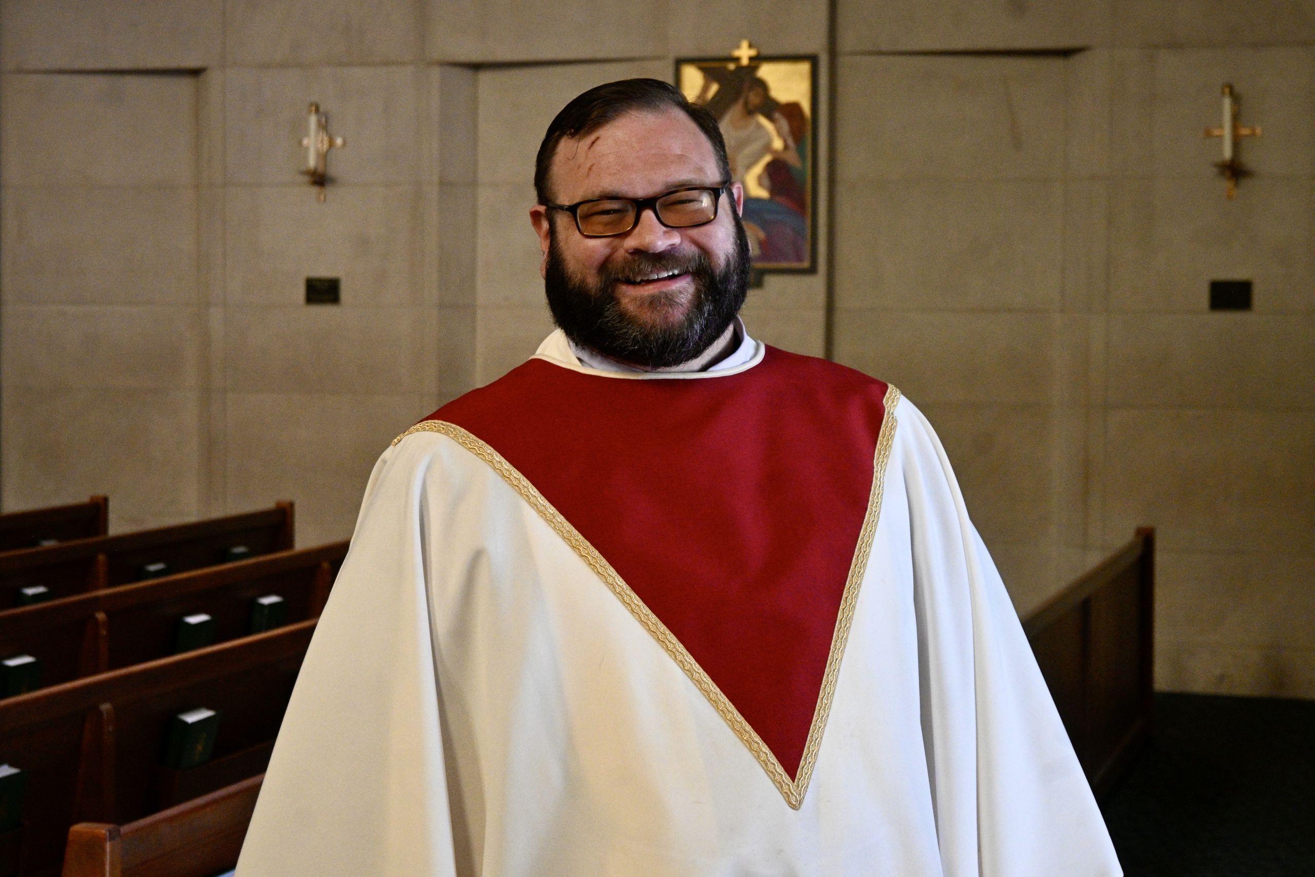 Smiling man in white liturgical robes with a red stole, standing in a church sanctuary with pews nearby.