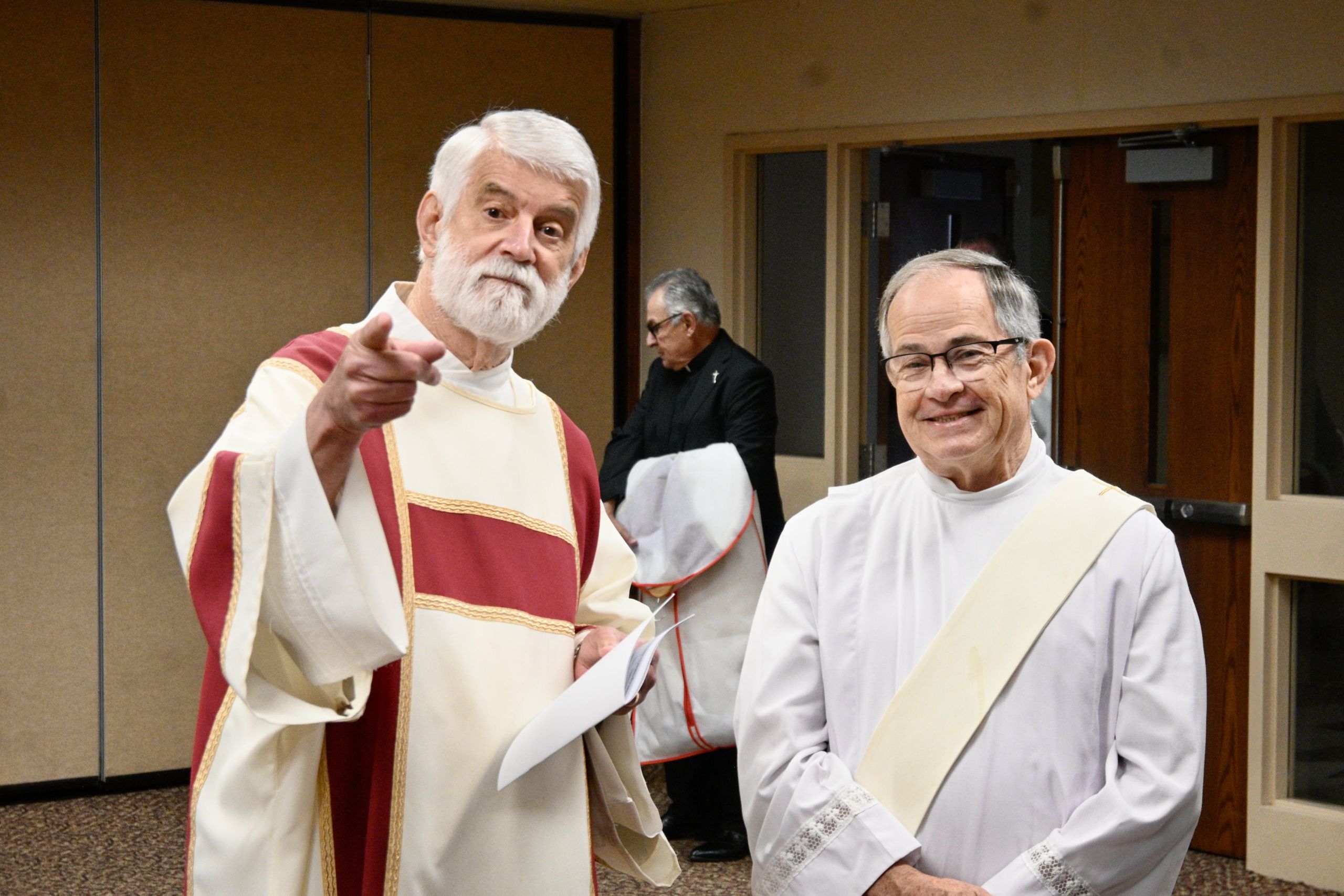 Two priests in cream and red liturgical vestments stand in a hallway; the bearded man points toward the camera while holding papers.