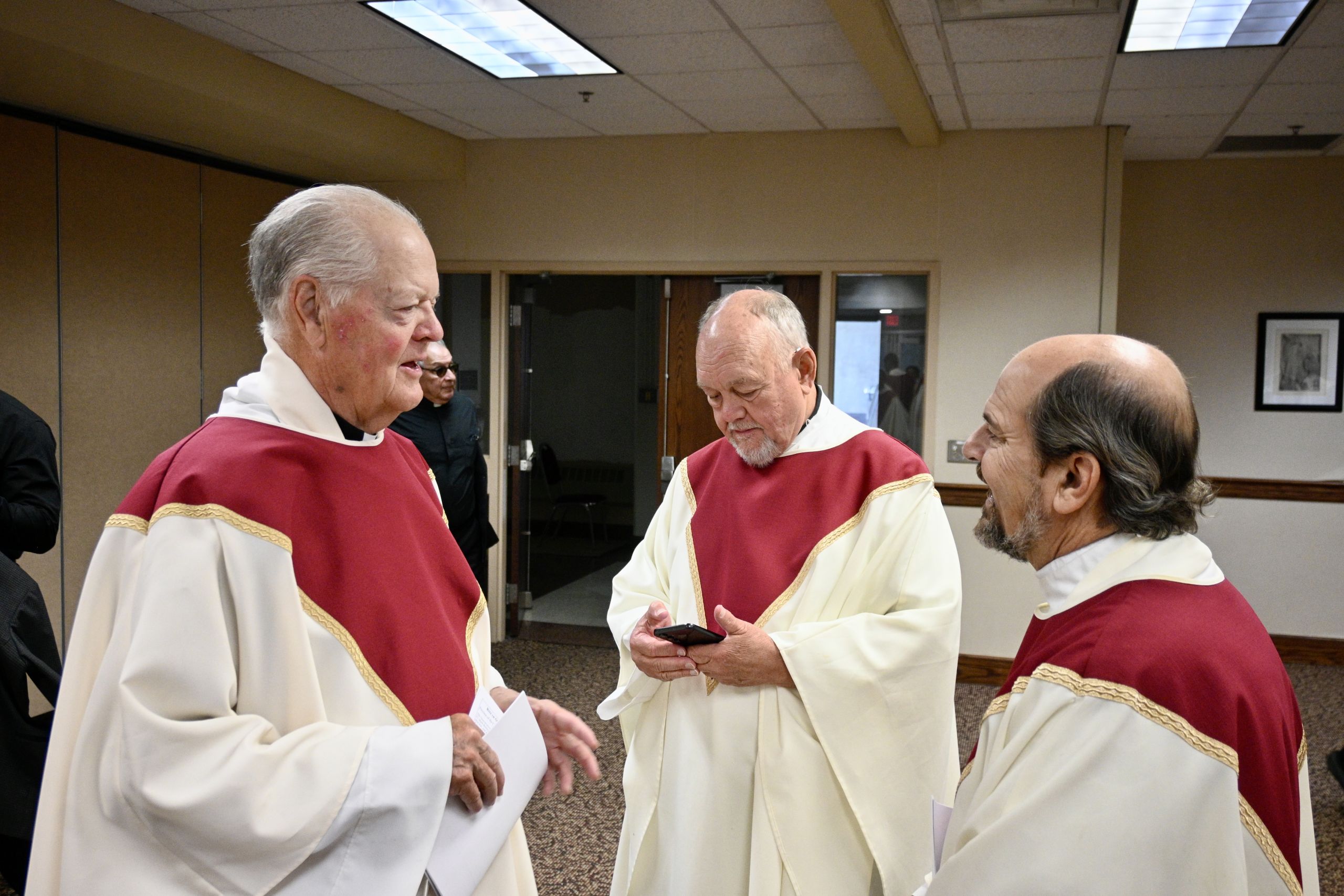 Three elderly men in cream and maroon choir robes stand in a room, one holding a paper while another checks a smartphone as they converse.