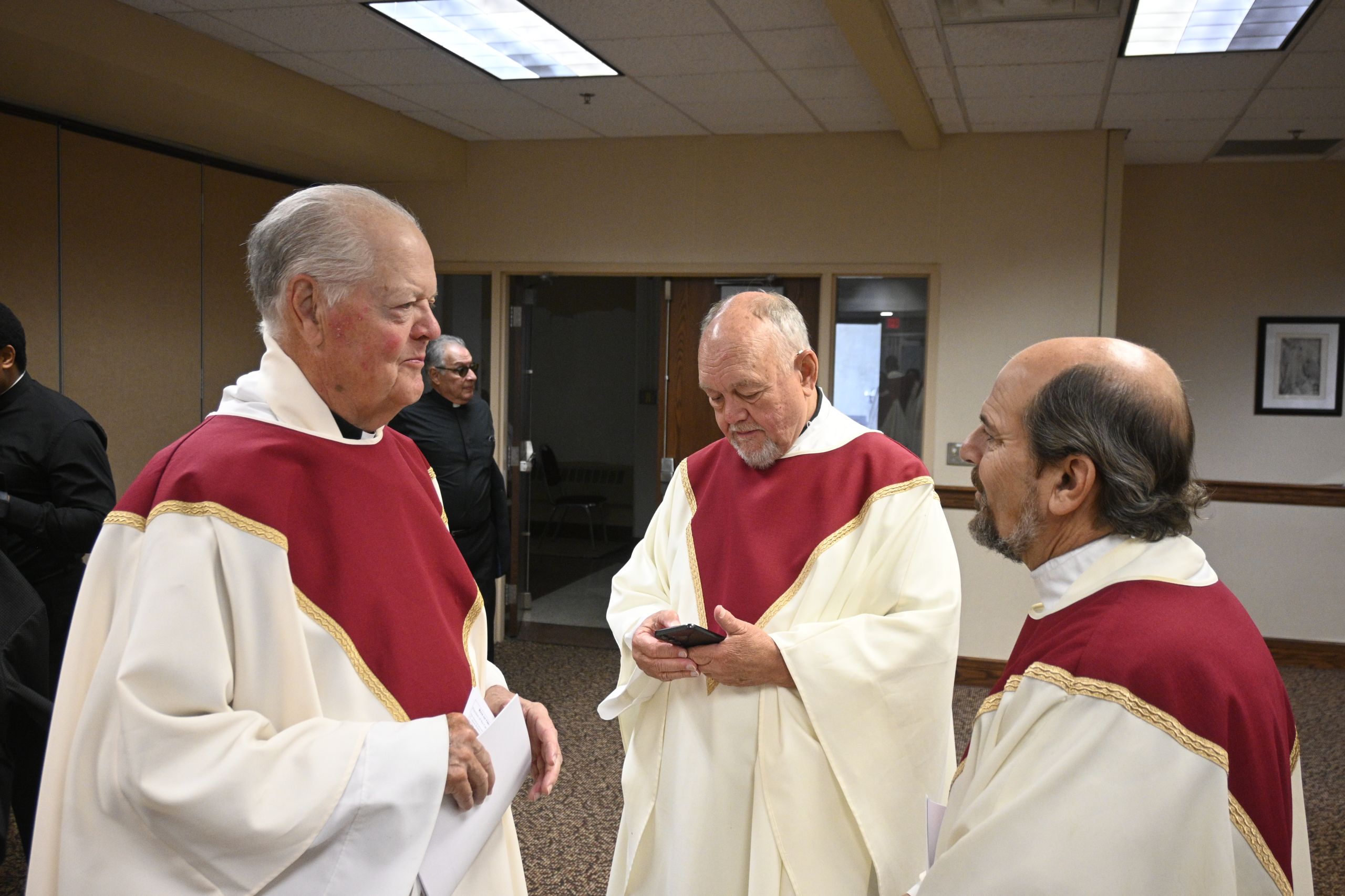 A group of elderly men in cream and maroon choir robes standing in a hallway, one man looking at a phone while others converse.