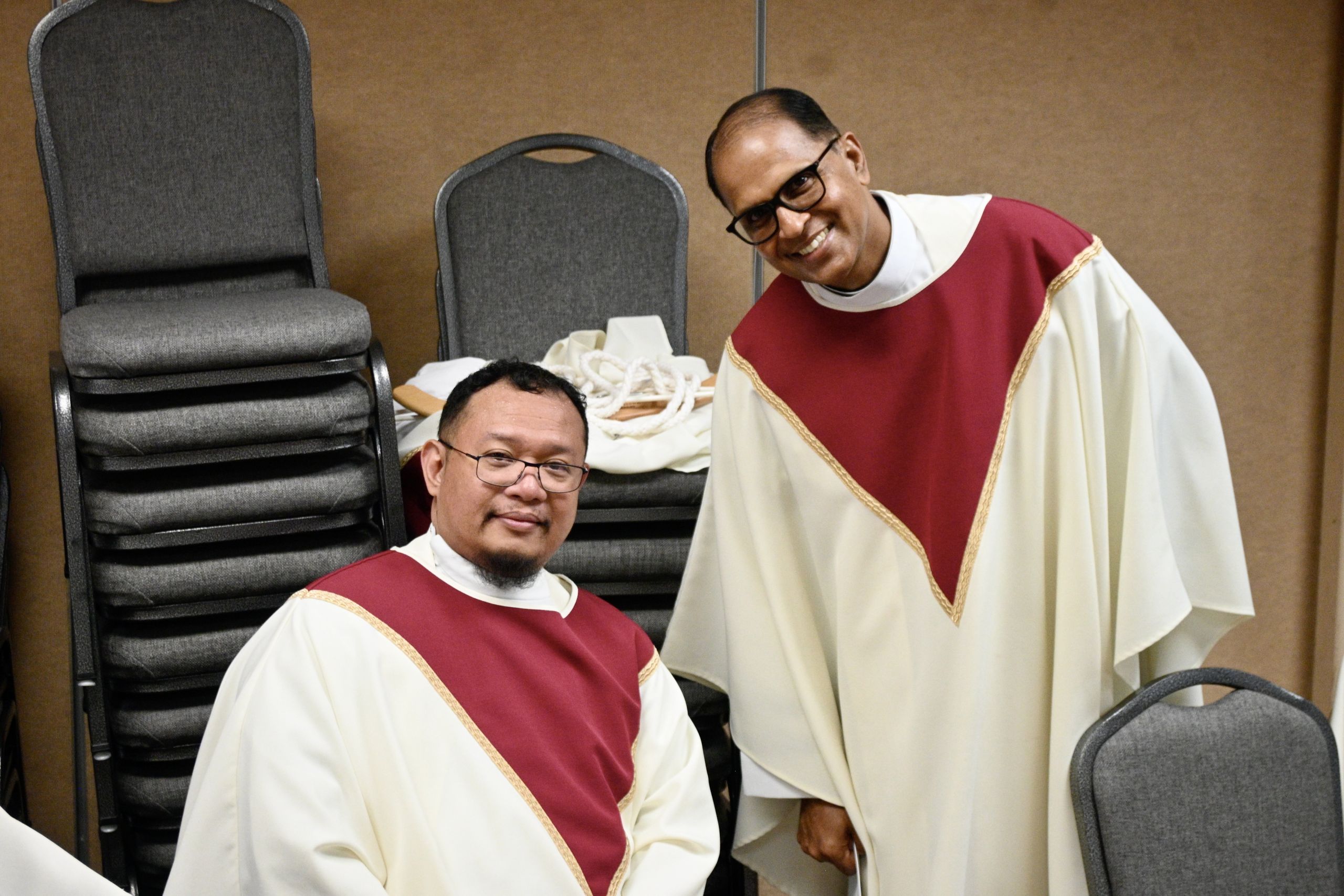 Two men in white and maroon choir robes posing and smiling beside stacked chairs in a room.