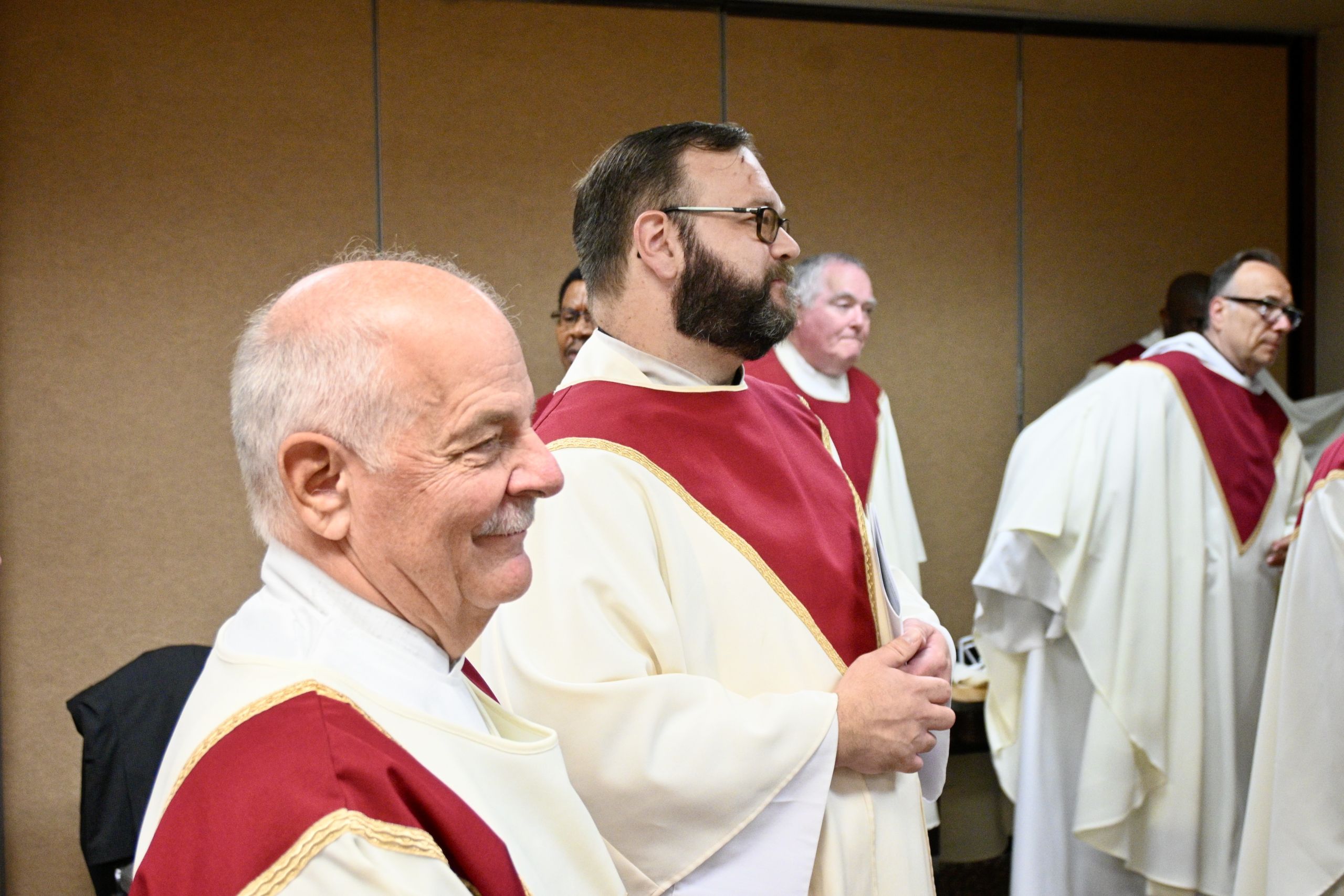 Clergy in cream and burgundy liturgical robes standing in a line indoors, hands folded.