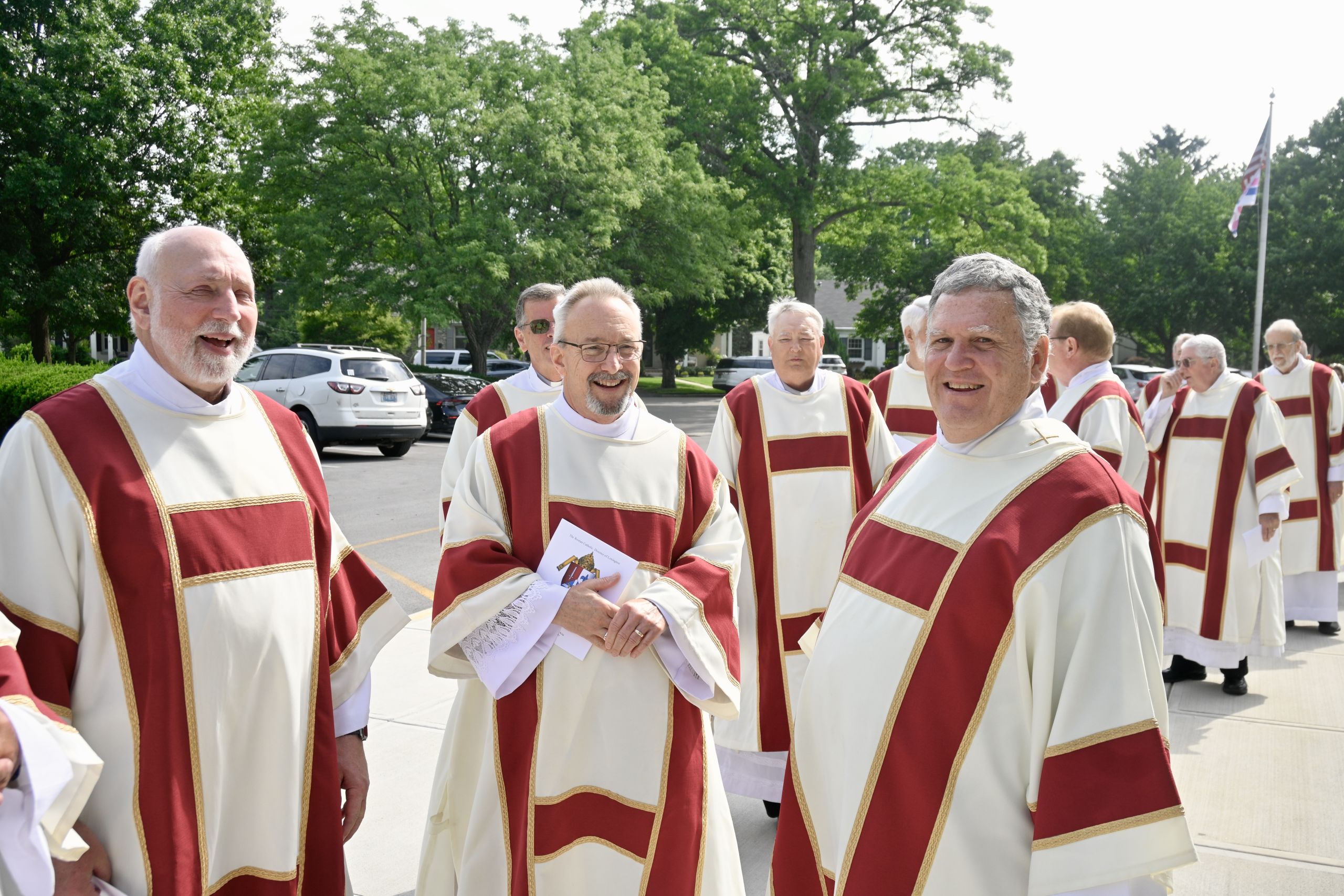 Group of clergy in white and red liturgical robes standing outdoors, smiling, with trees and parked cars in the background.