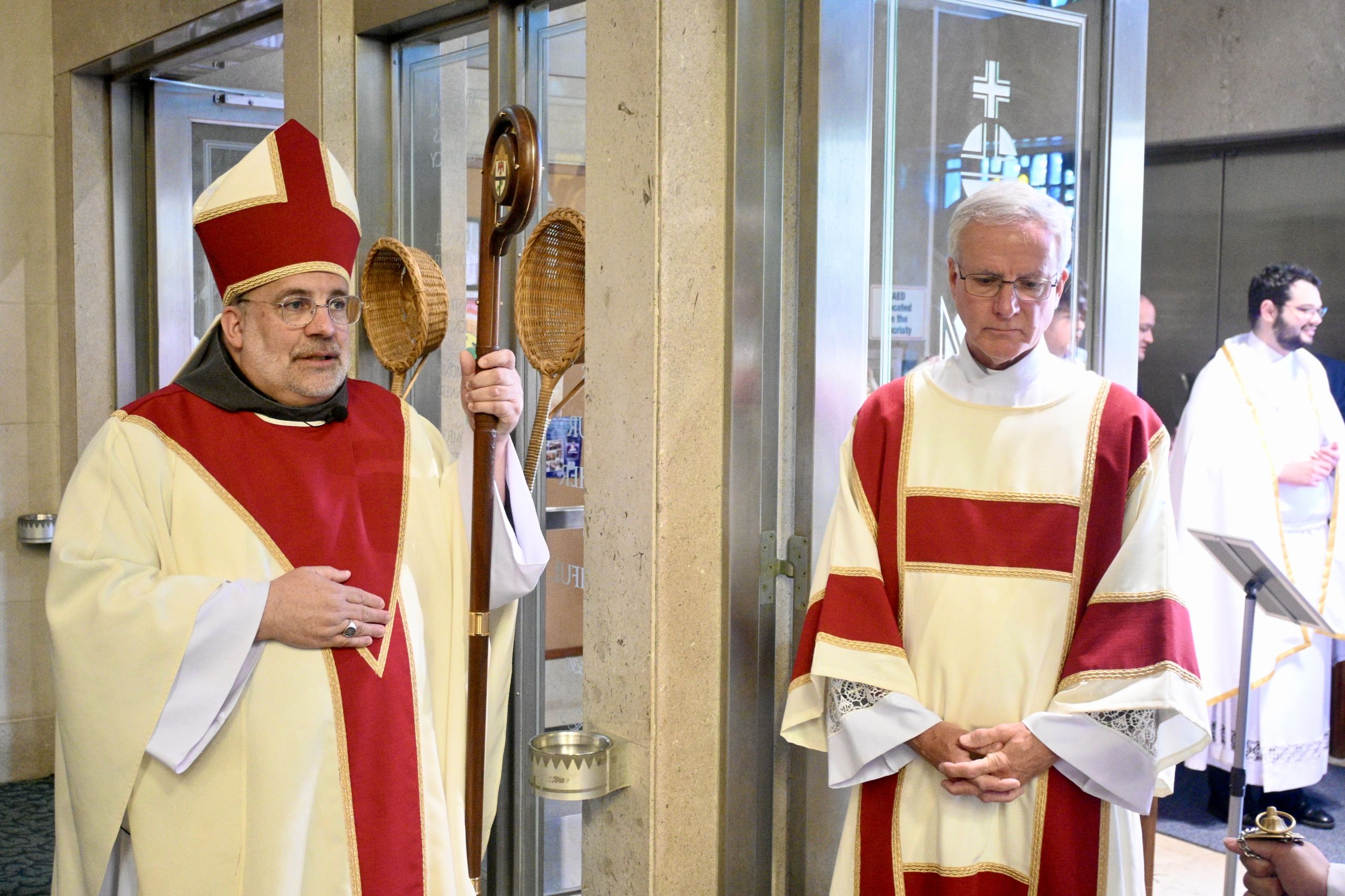 Two clergy in cream and red liturgical vestments stand in a doorway; the left figure holds a crozier and wears a tall red mitre.