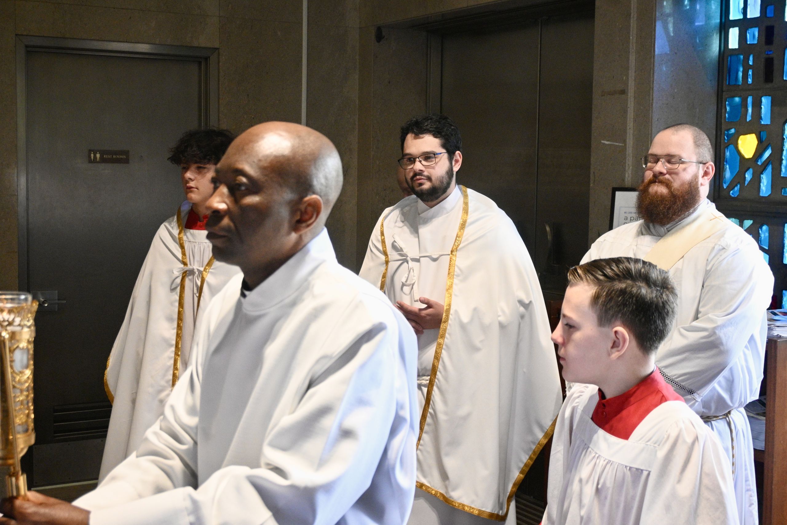 Group of men in white ceremonial robes with gold trim standing in a line indoors, likely part of a religious service.