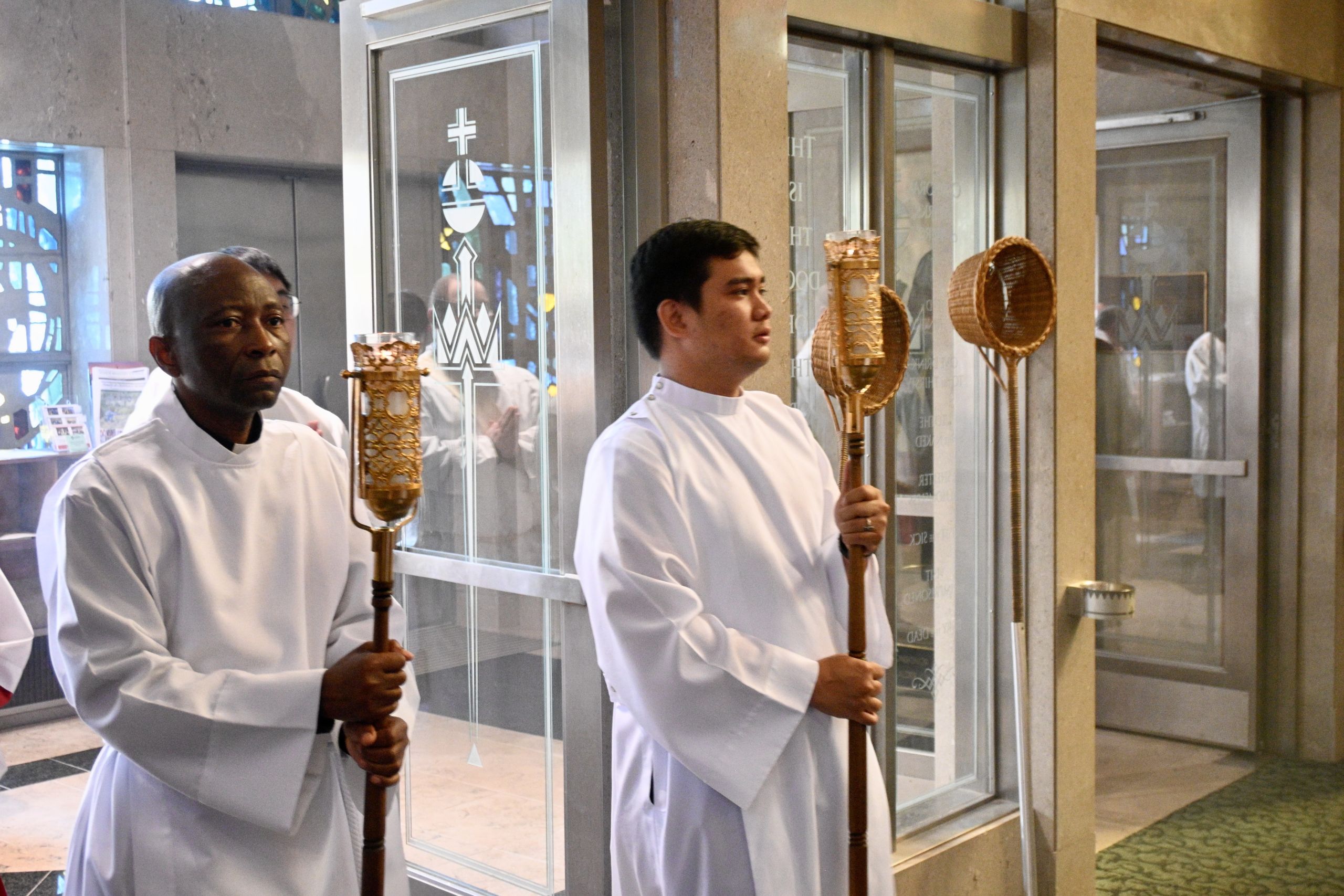 Two men in white liturgical robes stand holding tall ceremonial staffs inside a glass-walled lobby, with ornate golden reliquaries nearby.