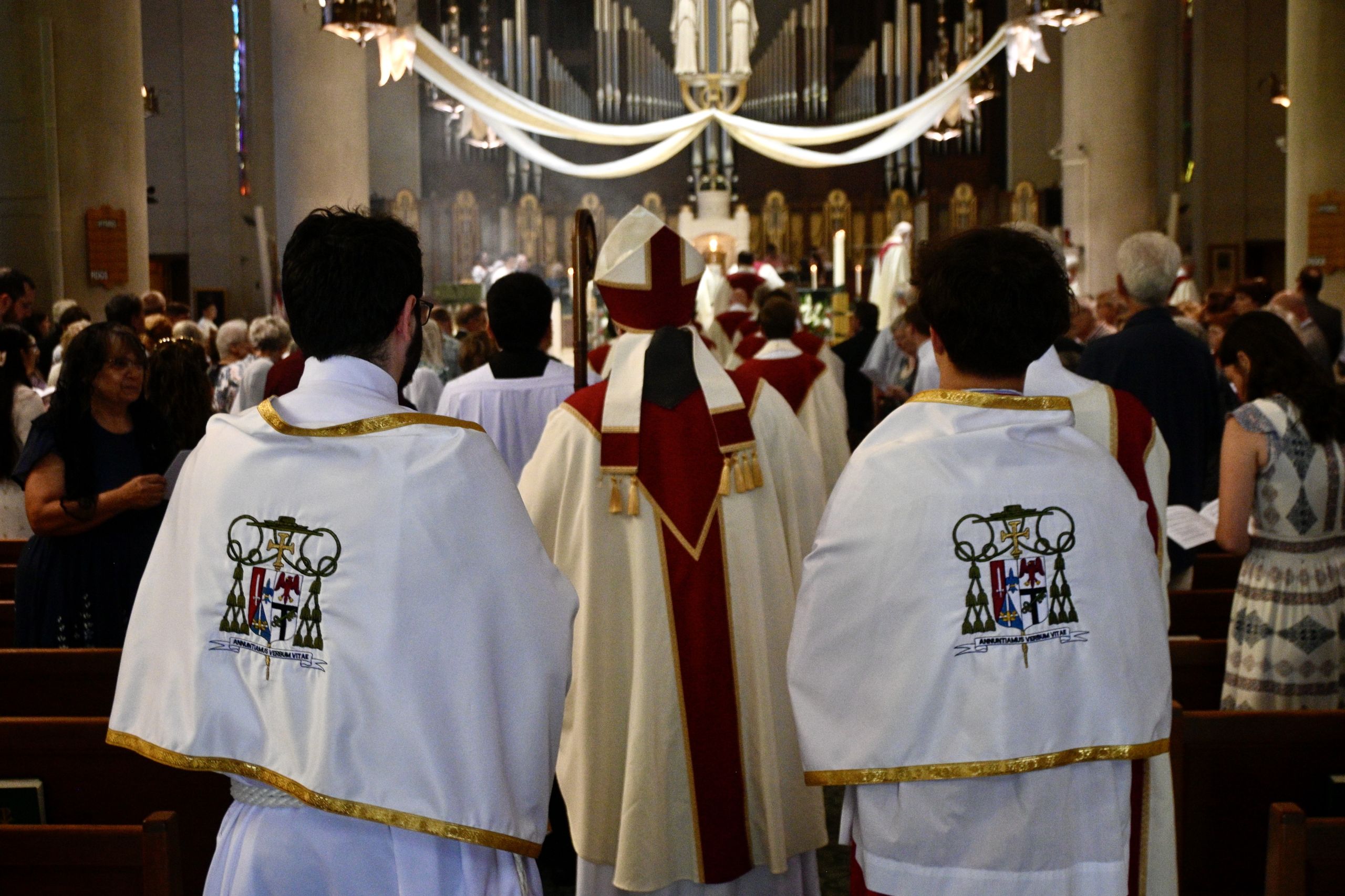Back view of clergy in white robes with gold trim, facing an altar during a church service; organ pipes and crowd in a dimly lit sanctuary behind them.