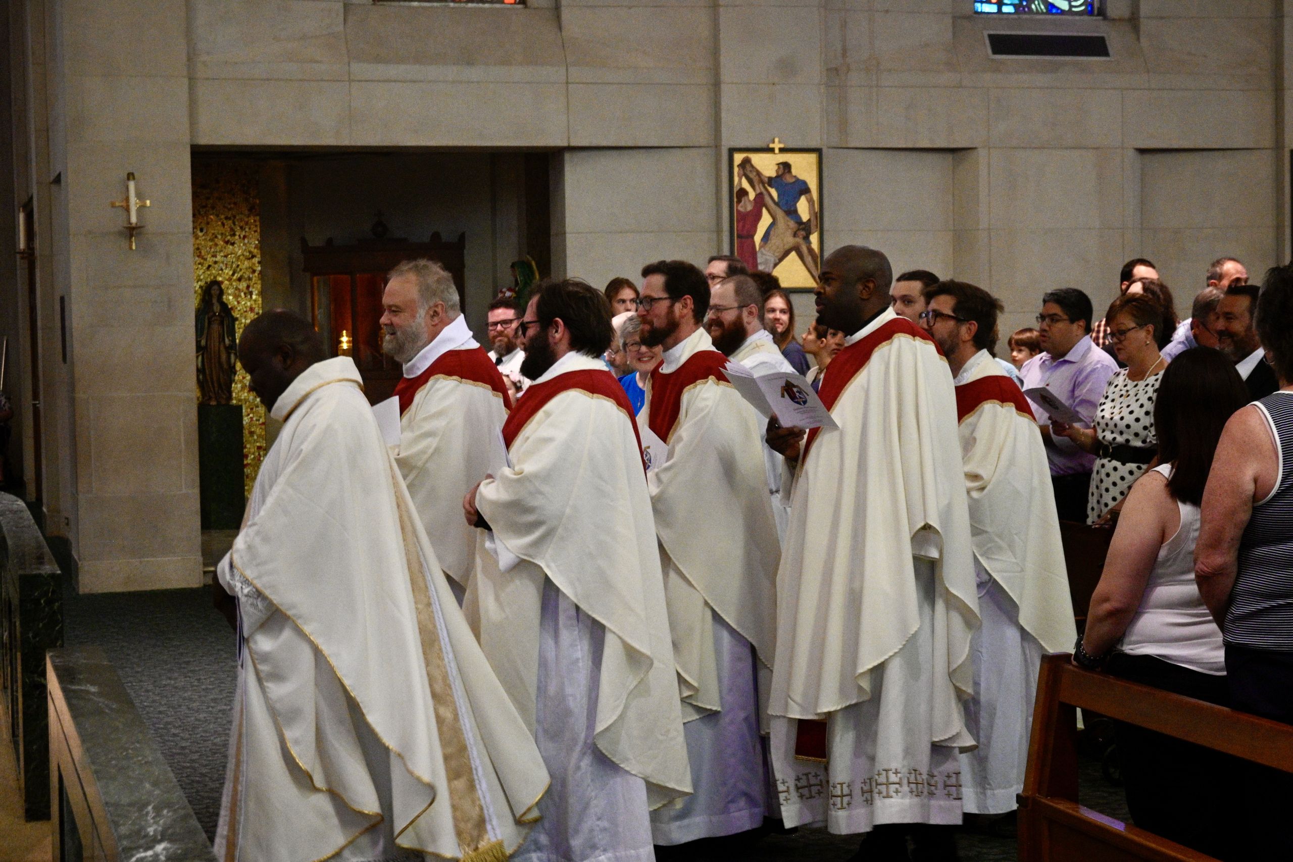 Line of Catholic priests in white vestments with red stoles walking in a church procession, followed by congregants standing along pews.
