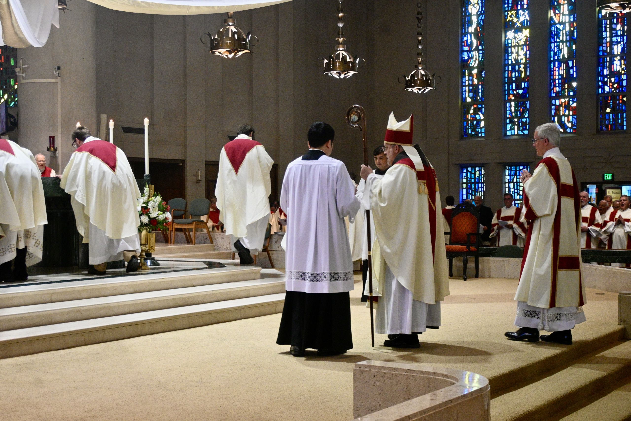 Clergy in white and red vestments lead a church ceremony on marble steps, bishop with crozier present in a dimly lit church.