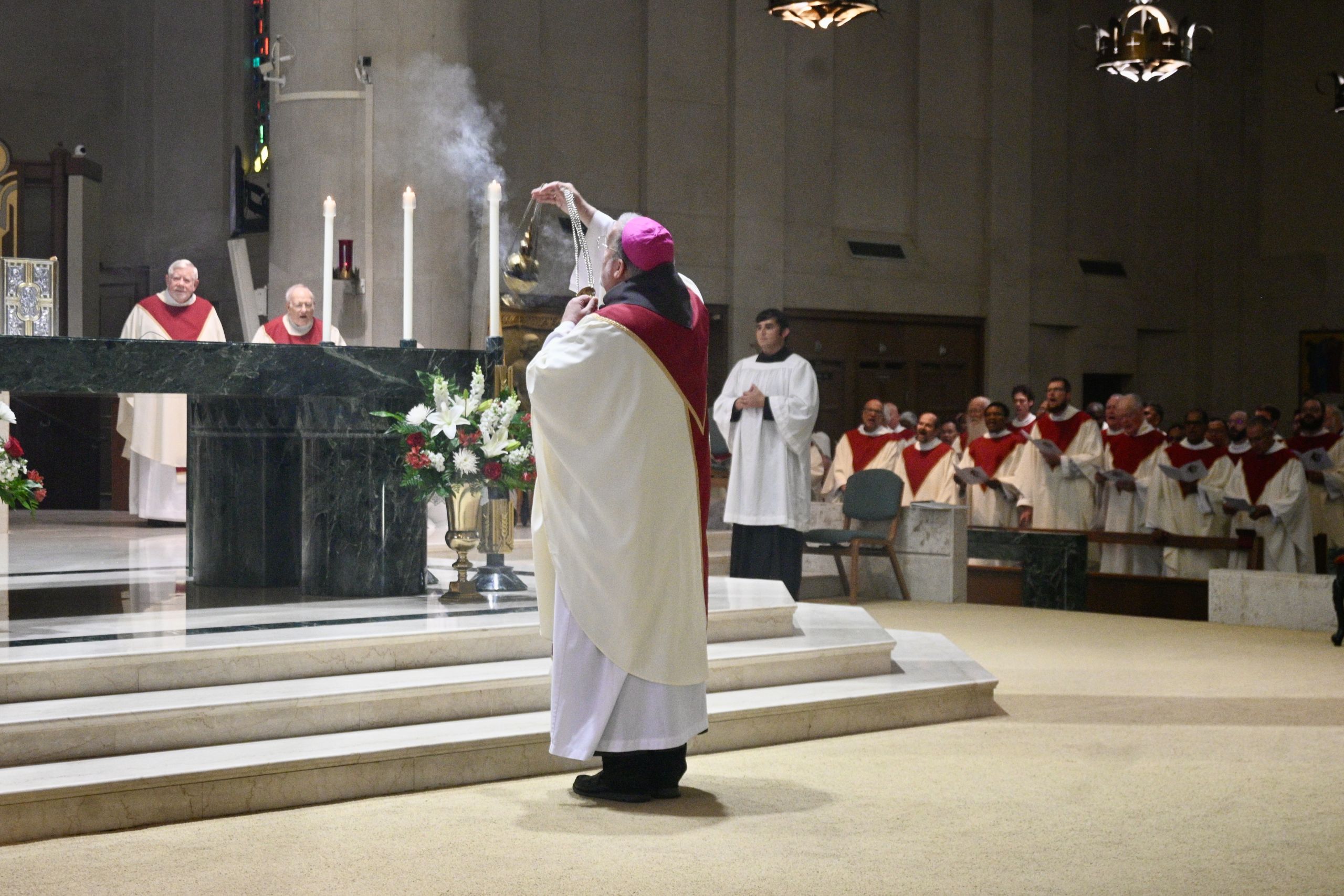 Bishop in pink zucchetto swings an incense censer over the altar while priests in red-and-white vestments look on.