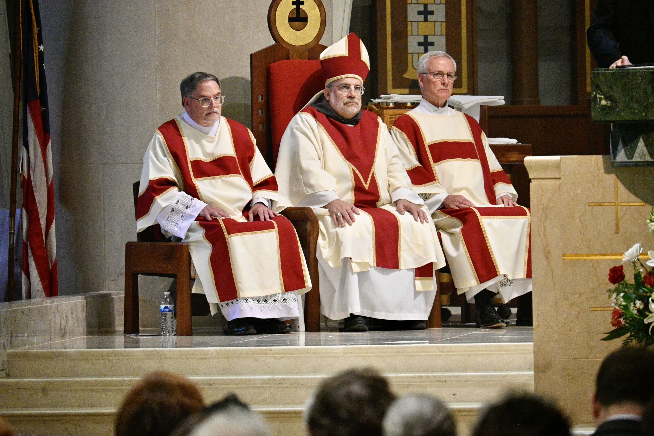 Three clergy in red-and-cream liturgical robes sit on a church dais during a ceremony, with a flag visible on the left.