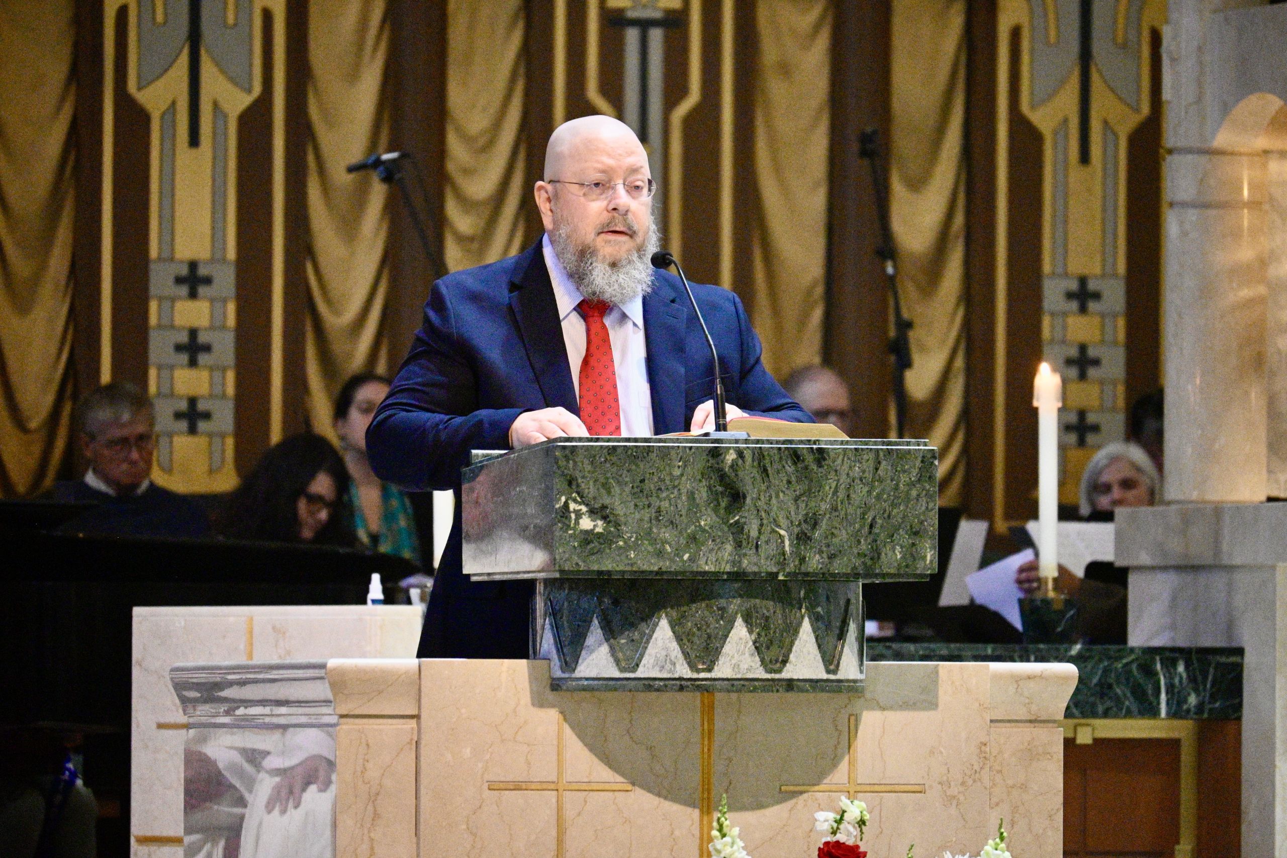 Bearded man in a navy suit and red tie speaks from a marble lectern in a church, microphone in front of him.