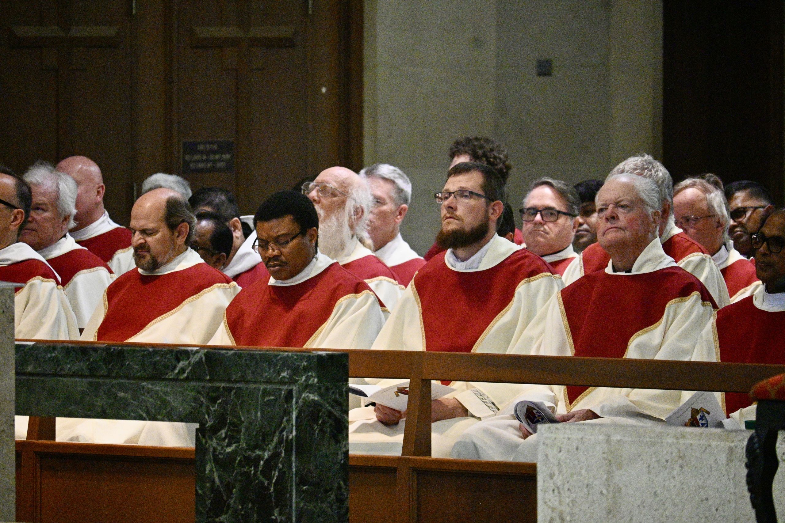 Clergy in red and white liturgical robes seated in a church pews during a ceremony.