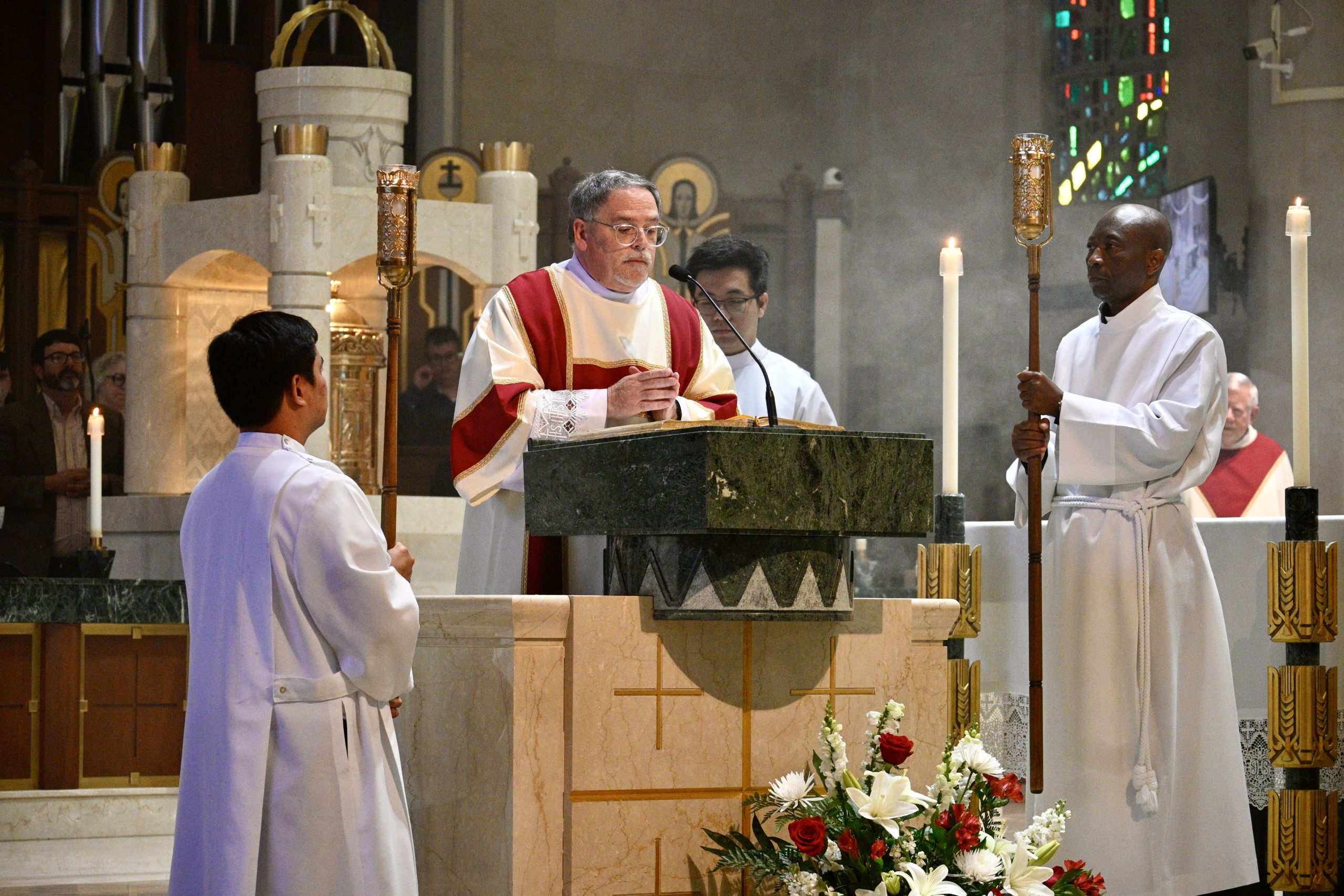 Priest in red and white vestments delivering a sermon at the marble altar, flanked by candle-bearing altar servers.