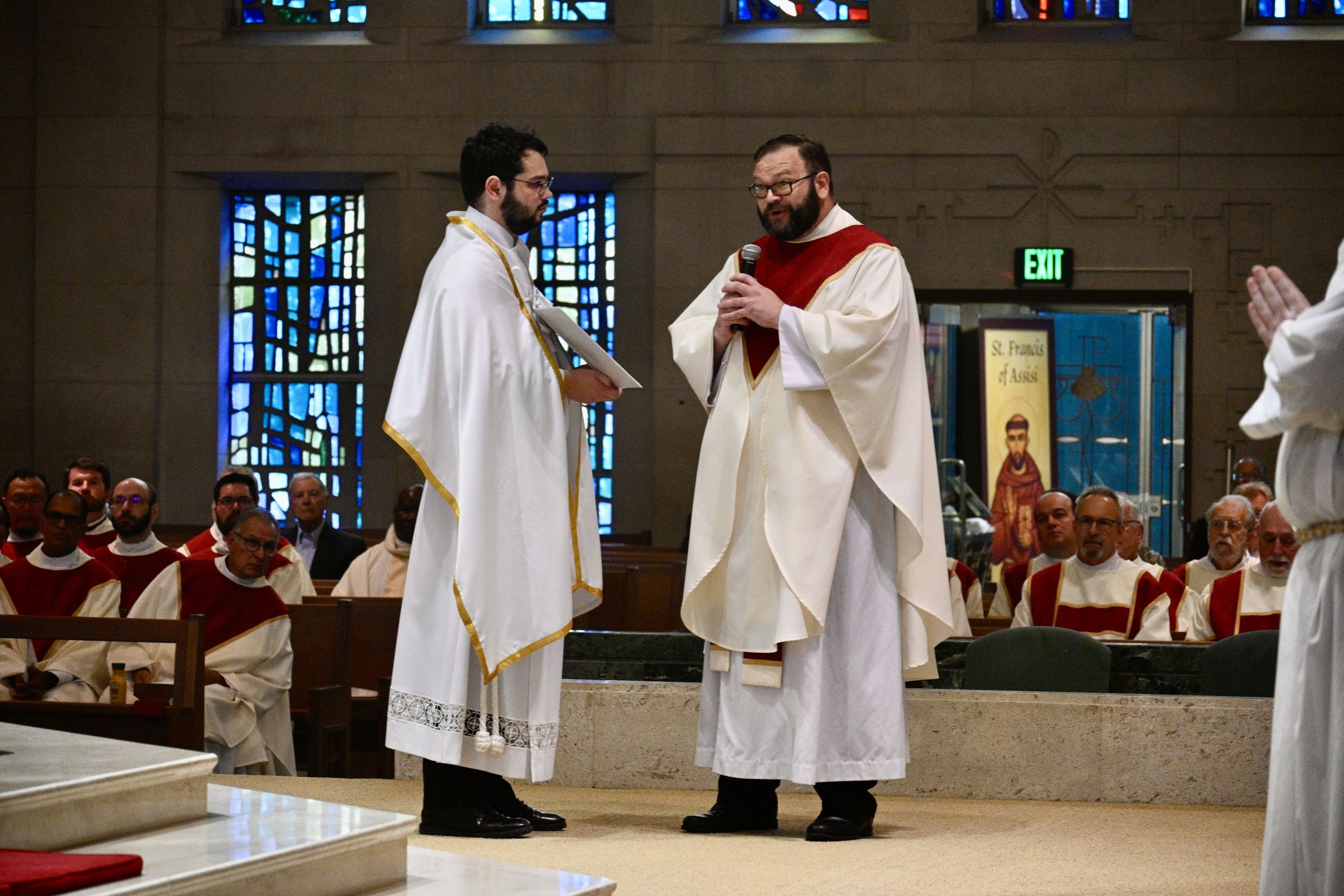 Two clergy in white robes with gold trim share a moment at a church ceremony; one holds papers while the other speaks into a microphone.