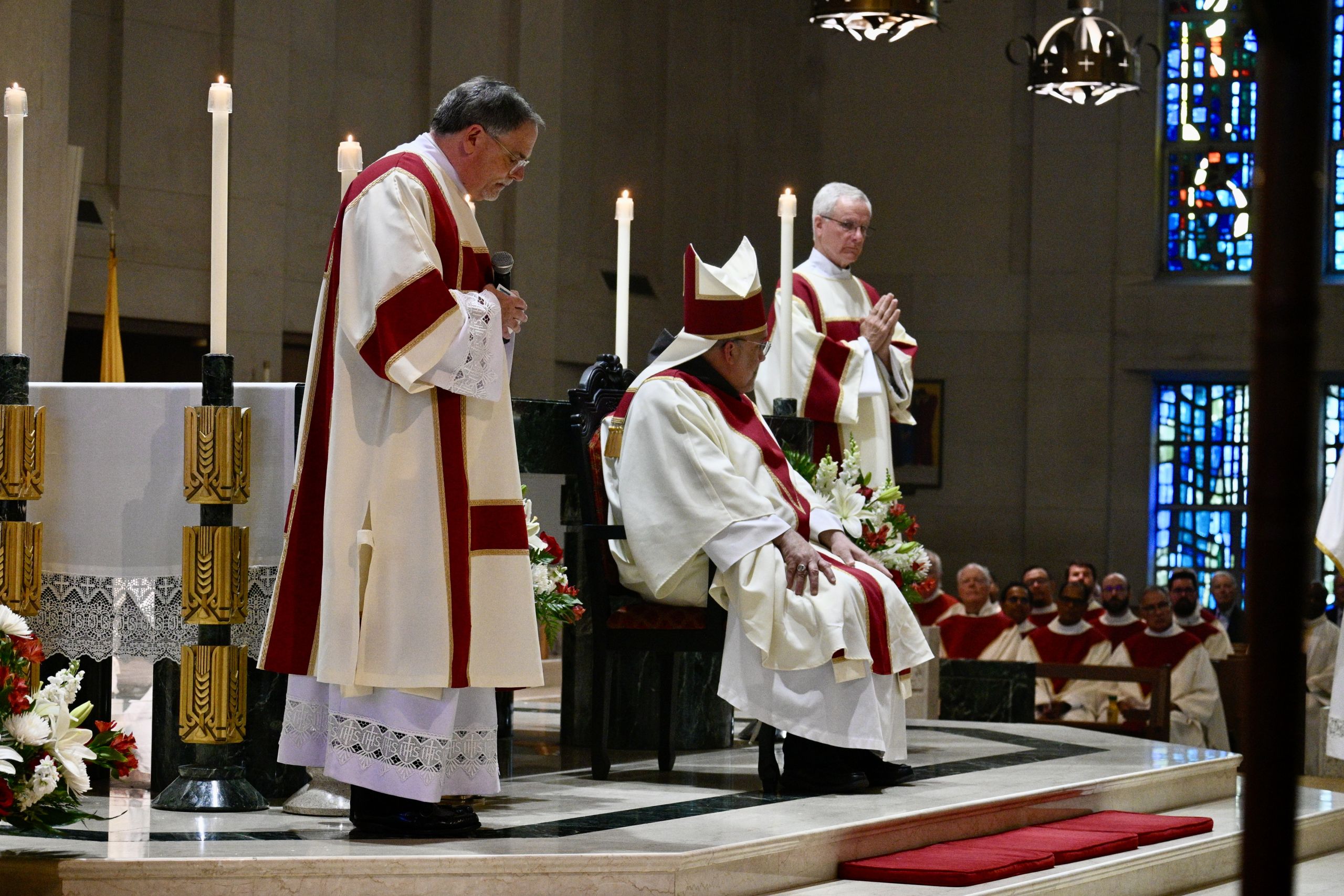 Catholic clergy in white and red vestments at the altar during a service with tall candles and floral arrangements nearby
