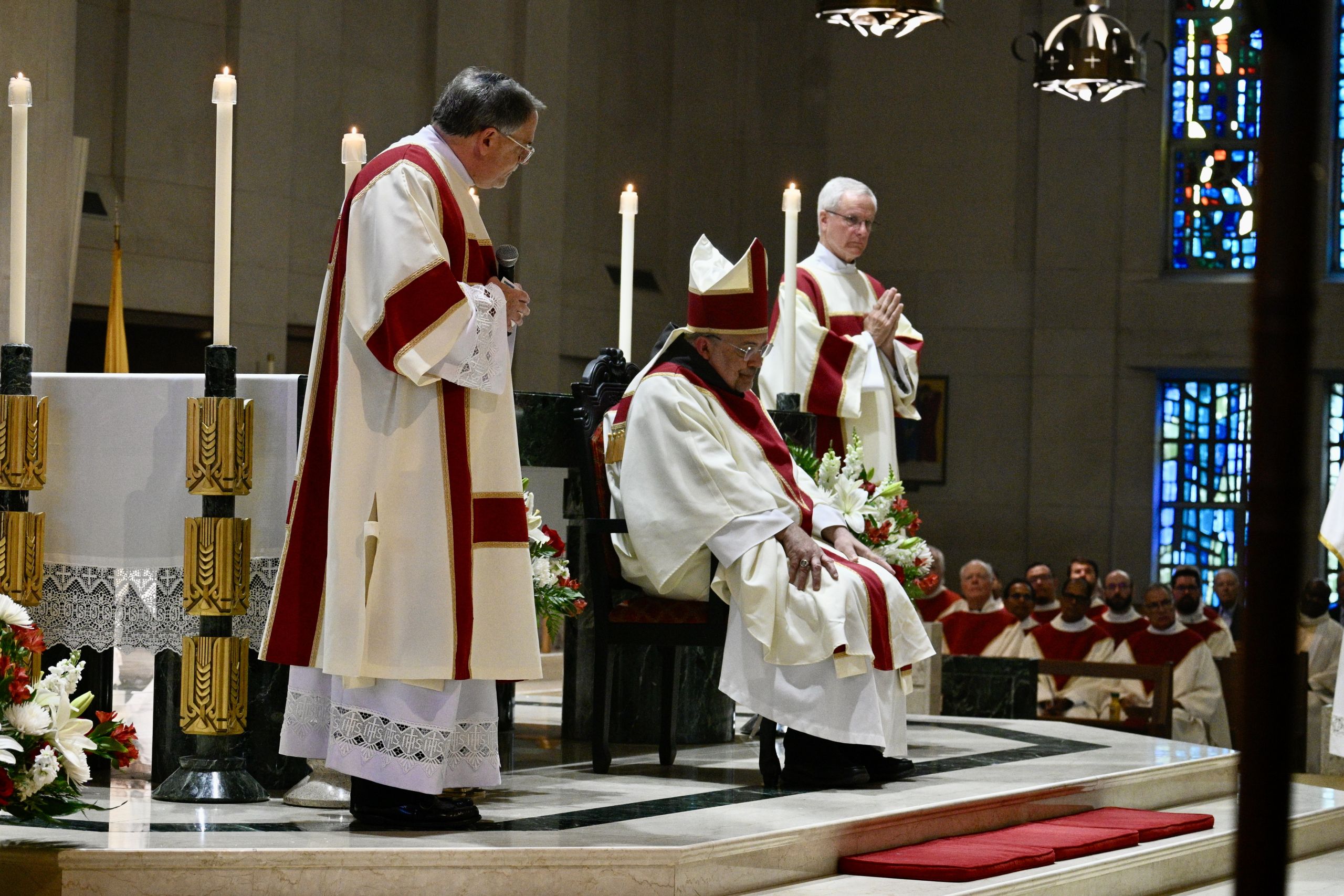 Catholic priests in red and white vestments conduct a ceremony at the altar with tall lit candles nearby in a church.