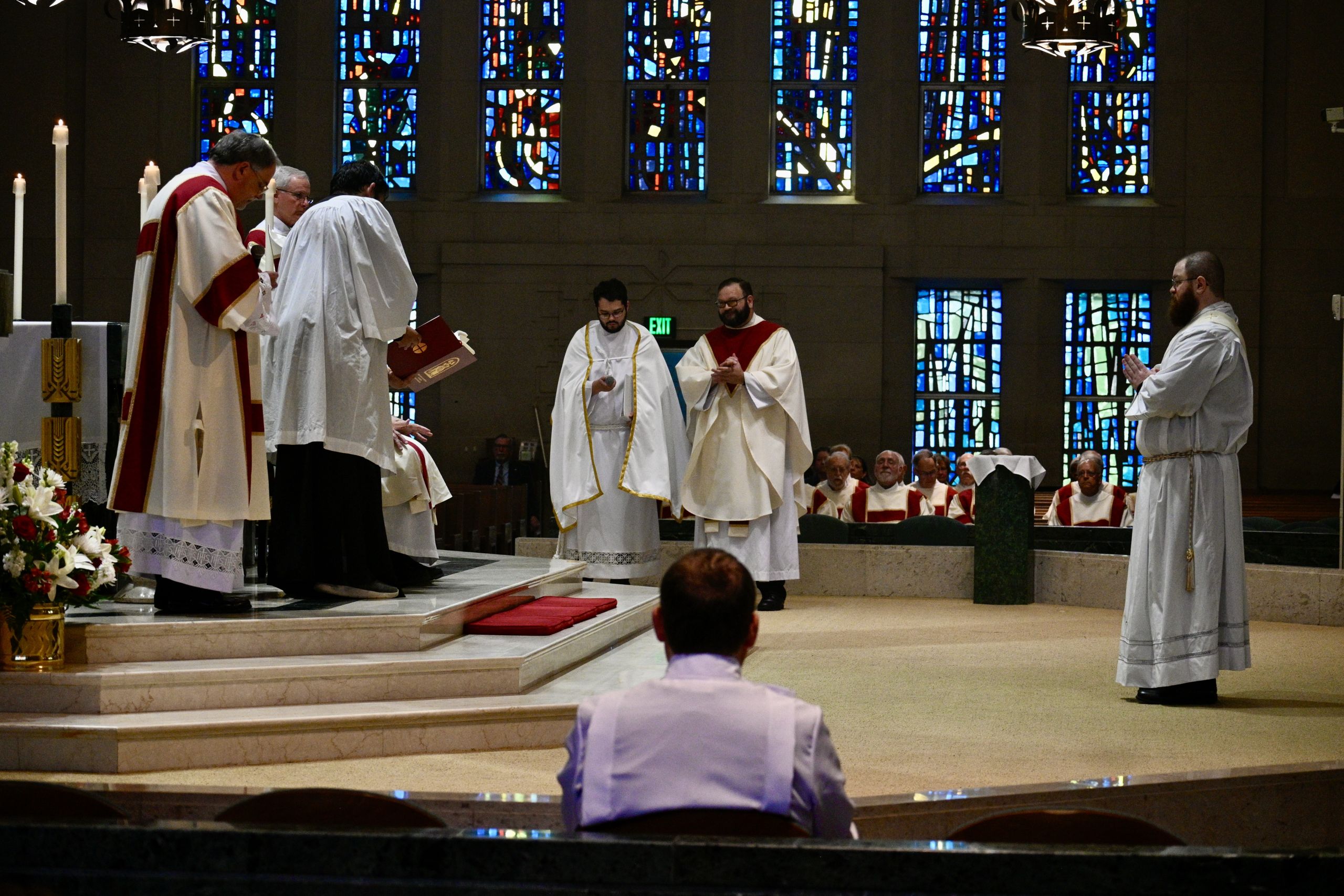 Priests in white robes with red stoles stand at the altar during a church service, colorful stained glass windows behind them.