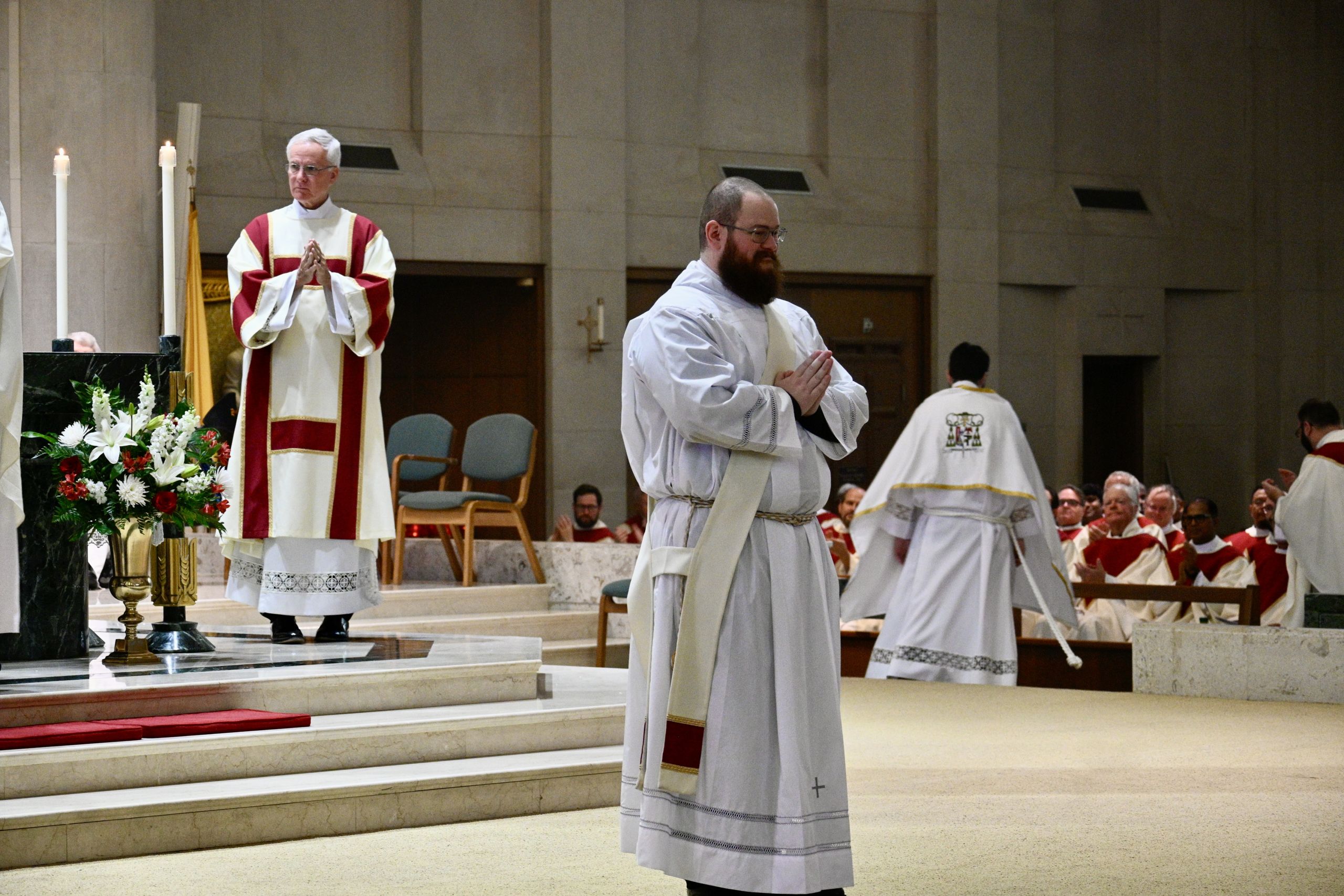 Priest in white and red robes standing at altar with hands in prayer during a church service, floral arrangement nearby.