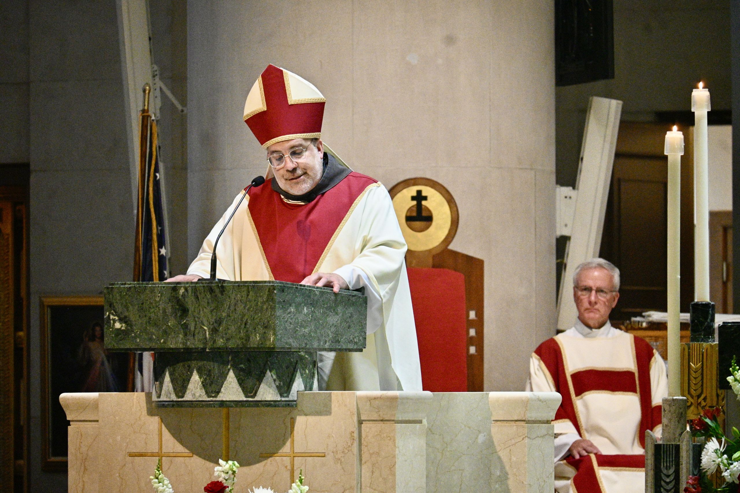 A Catholic bishop delivering a sermon at a marble pulpit during mass, wearing red and white vestments and a mitre.