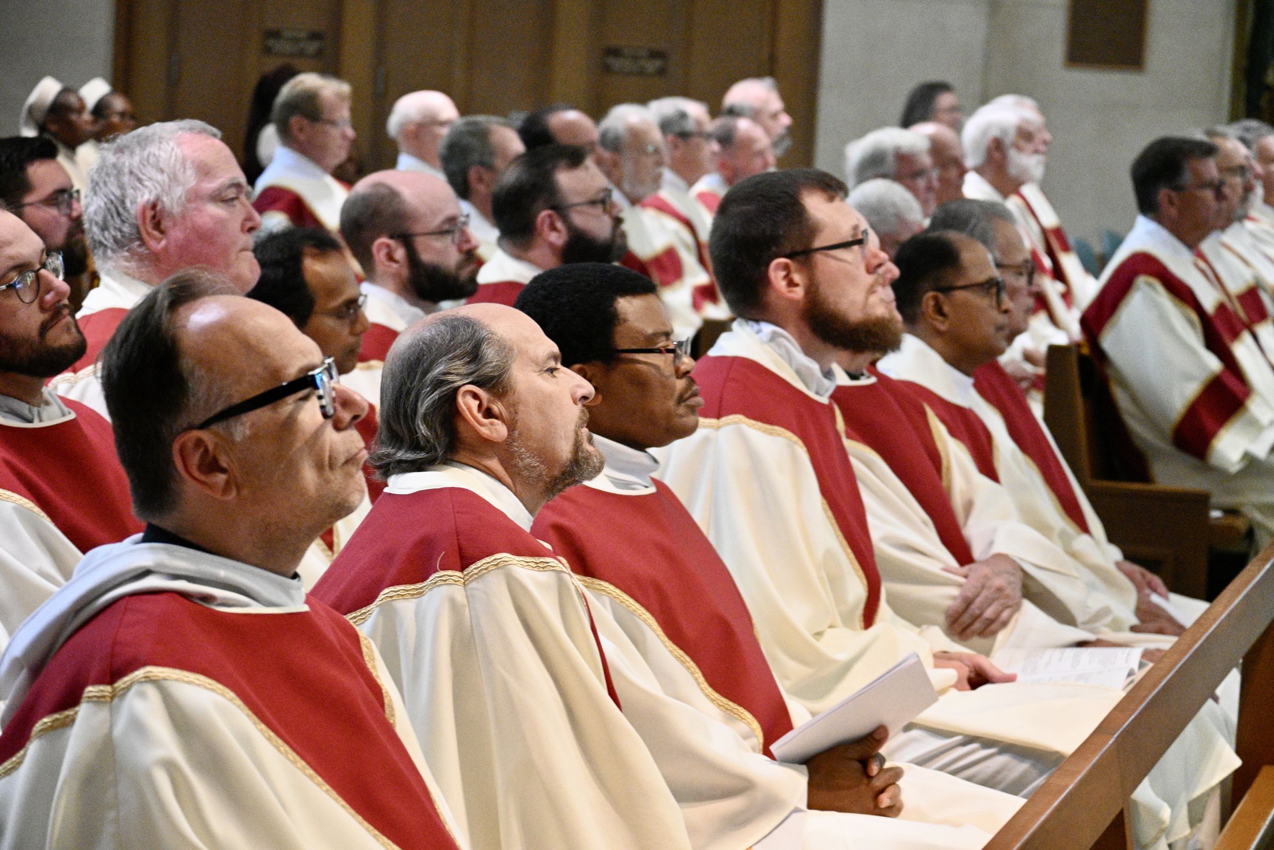 Group of clergy in white and red vestments seated in church pews during a ceremony.