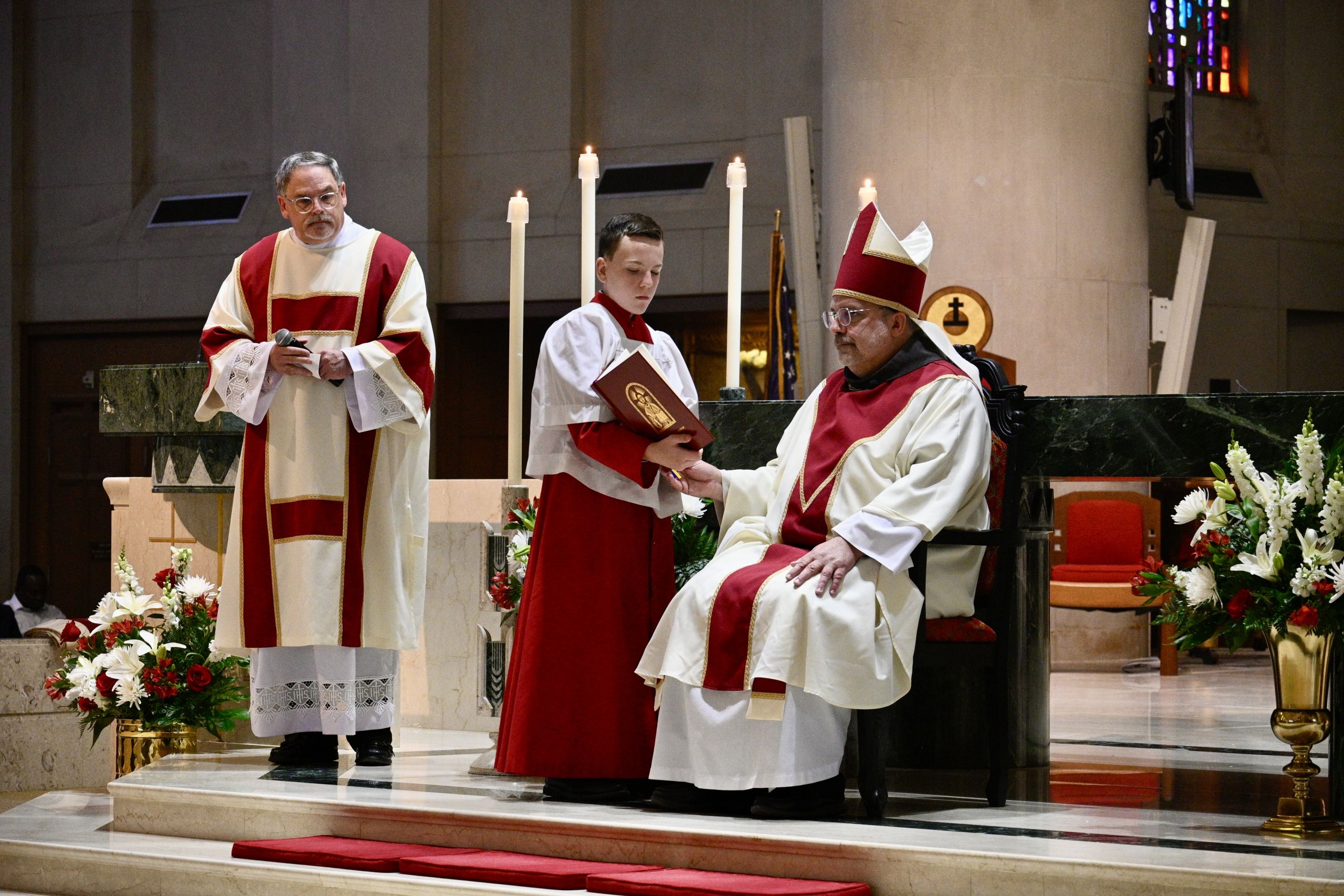 Clergymen in red and white liturgical robes at an altar with candles and flowers inside a church.